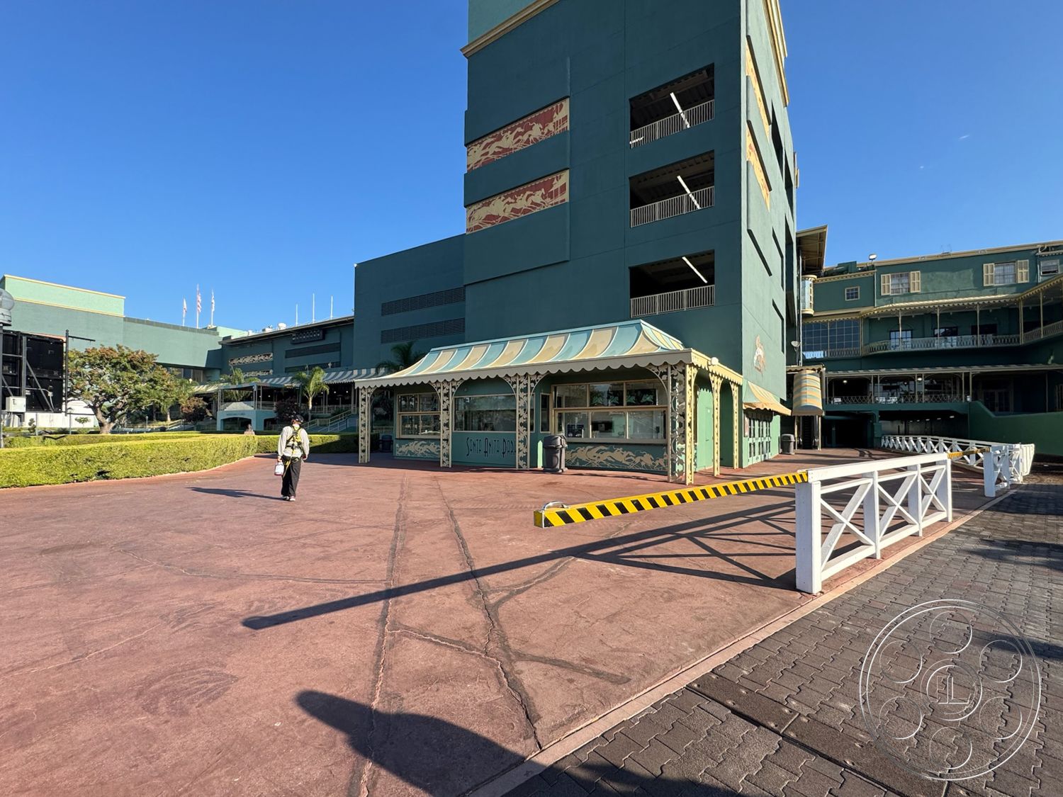 Arcadia Race Track - exterior, urban setting, paved walkway, modern architectural style, commercial property, concrete parking lot, sunlit outdoor space, landscaped garden with shrubs, asphalt access road, multistory building with green exterior, decorative entryway awning, ornate building facade, horizontal band of windows, striped caution barrier, multiplex structure, bilateral symmetry in design