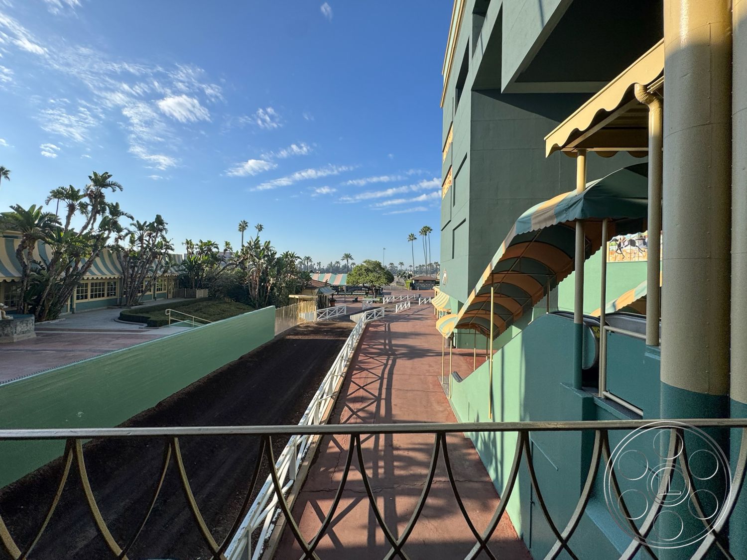 Arcadia Race Track - exterior, tropical landscaping, concrete walkways, urban backdrop, wrought iron details, stucco wall finish, decorative metal railing, arched window frames, green painted concrete walls, striped canvas awnings, paved racetrack, palm trees lining the area, amphitheater-style seating area, open-air horse racing venue, sunshine casting shadows, colorful exterior accents