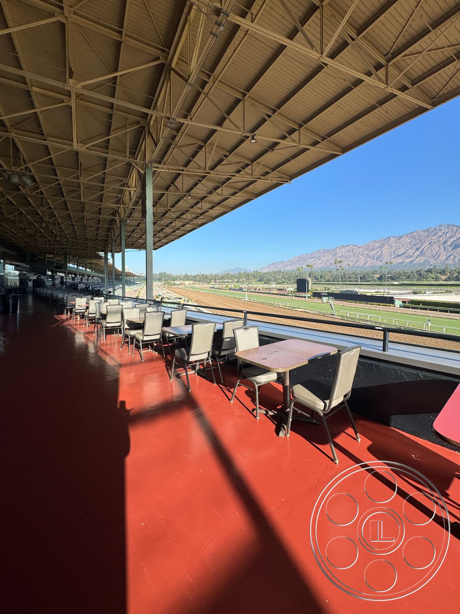 Arcadia Race Track - mountain backdrop, exterior seating area, sunlit terrace, wood beam ceiling, concrete support columns, spacious patio area, metal frame roof structure, red epoxy-coated floor, outdoor viewing deck, racing track view, commercial outdoor seating, aluminum railing system, covered outdoor space, weather-resistant furniture, viewing area for events