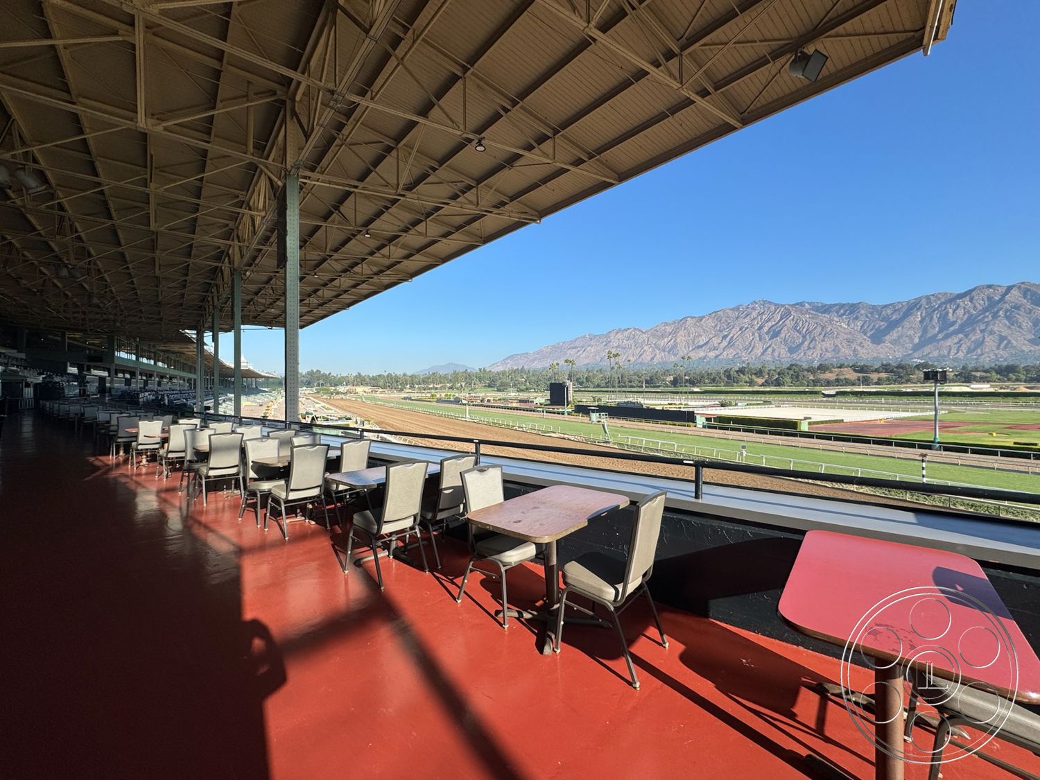 Arcadia Race Track - landscaped surroundings, outdoor dining area, mountain view, steel support columns, clear blue sky backdrop, exterior grandstand structure, metal roof canopy, red epoxy-coated concrete flooring, racing track observation deck, commercial restaurant seating, open-air seating arrangement, horse racing venue, urban racecourse setting