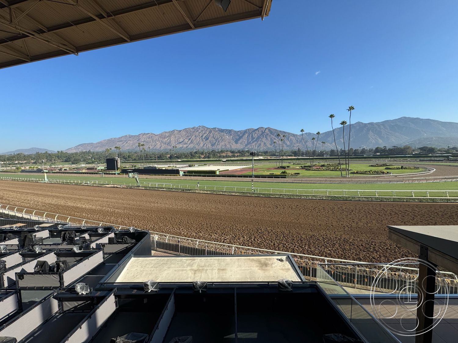 Arcadia Race Track - exterior, metal railings, paved walkway, open sky, sunny outdoor environment, mountain backdrop view, palm trees in foreground, concrete horse racing track, dirt racetrack surface, landscaped greens surrounding racetrack, grandstand viewing area, expanded horse paddock area, large video screen for race viewing, spectator seating area, lush grass landscape, city skyline in distance