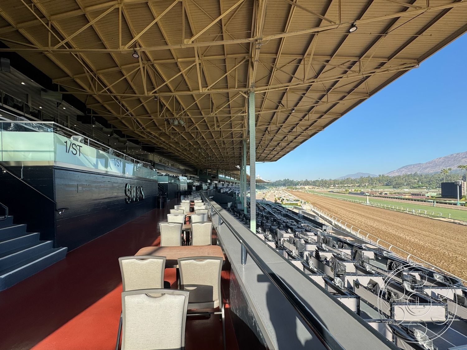 Arcadia Race Track - concrete flooring, glass railing, open-air seating, modern architectural style, mountain backdrop, wood beam ceiling, metal support columns, exterior grandstand structure, red polished concrete floor, outdoor viewing area, restaurant-style tables and chairs, panoramic racecourse view, urban racetrack setting, spacious outdoor terrace