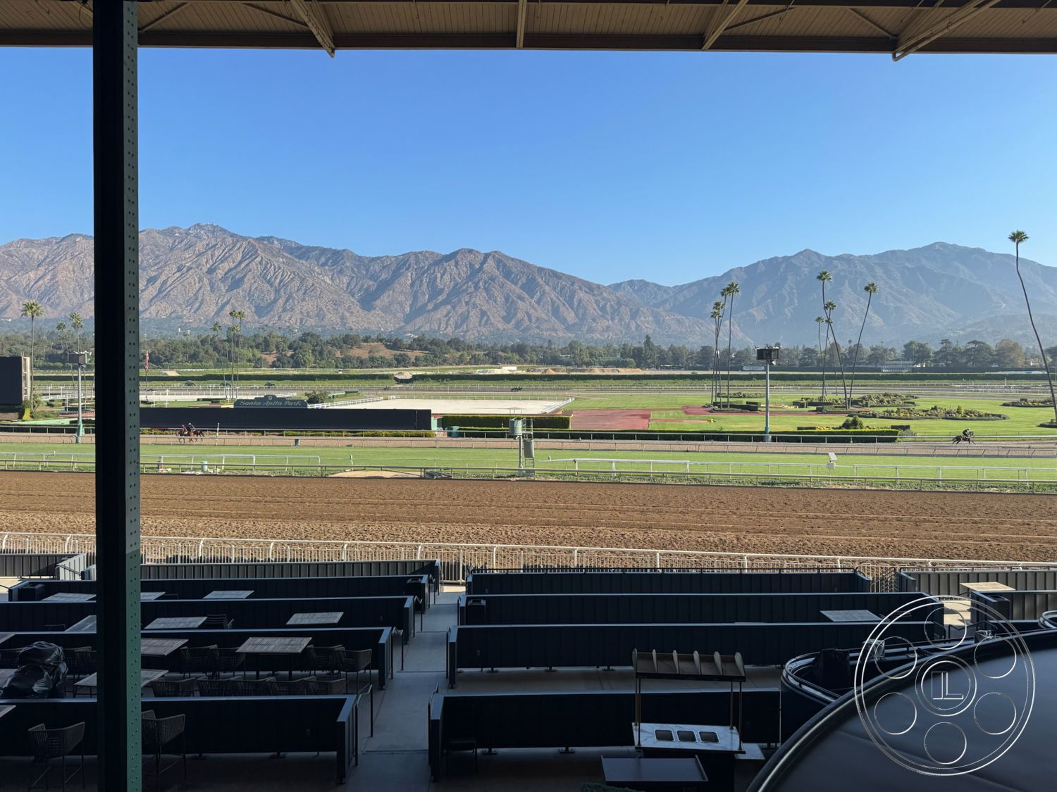 Arcadia Race Track - concrete flooring, wooden picnic tables, outdoor event space, landscaped grounds, steel support beams, sunny outdoor environment, aluminum railings, desert mountain backdrop, panoramic mountain view, exterior grandstand structure, open-air seating areas, landscaped horse racing track, paved horse path, bleacher-style seating, spacious viewing deck, desert climate, modern sports complex, palm trees in the vicinity, gray steel awning, rectangular seating layout
