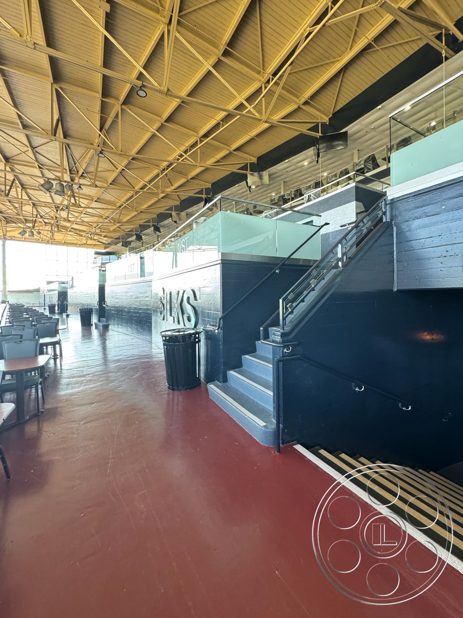 Arcadia Race Track - glass railing, modern interior, public space, dark wood accent wall, open-concept layout, indoor seating area, exposed wood ceiling beams, steel stair railings, industrial architectural style, yellow painted ceiling panels, concrete stair treads, red epoxy floor, restaurant setting, lively atmosphere