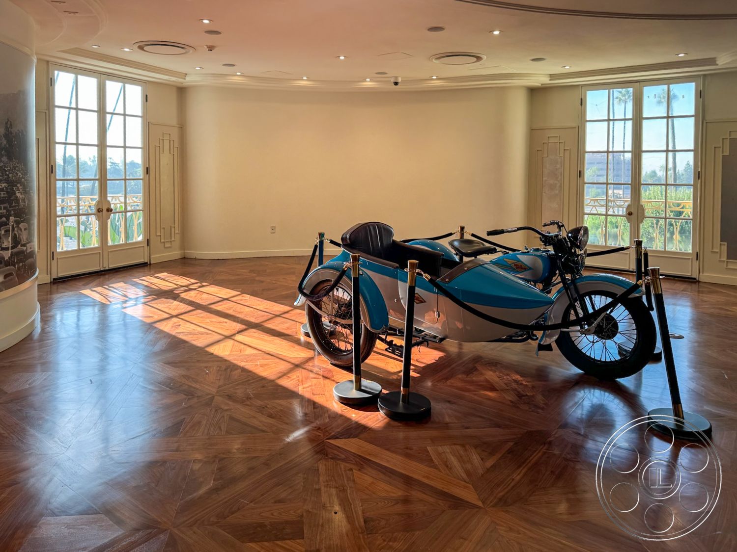Arcadia Race Track - exposed light fixtures, interior hardwood herringbone flooring, contemporary white painted walls, double French doors with brass hardware, decorative ceiling details, art deco design elements, large windows with view of palm trees, open spacious room, modern motorcycle with sidecar displayed, gold stanchions with black ropes for display, sunlit room with shadows on the floor, elegant gallery-like atmosphere