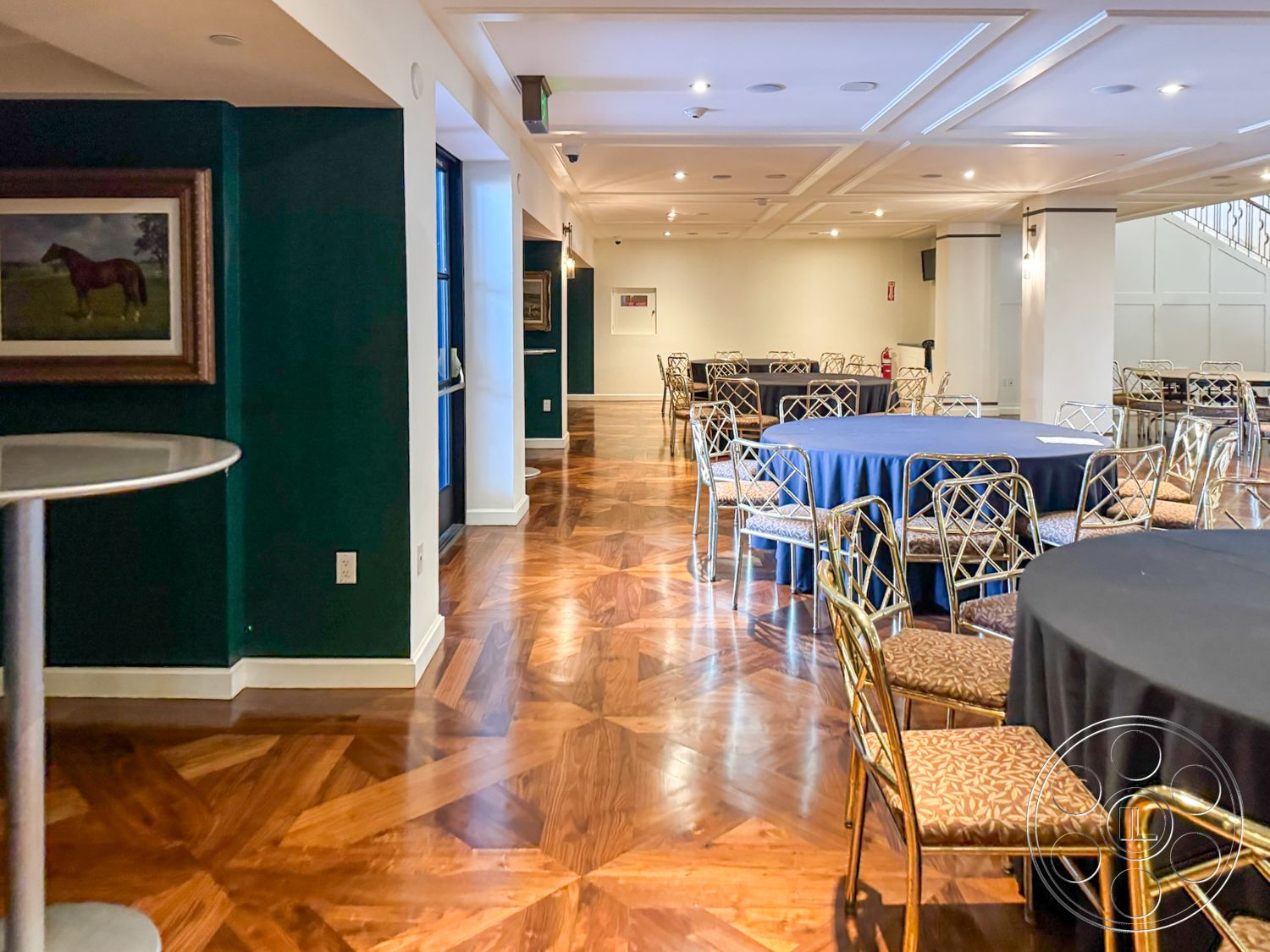 Arcadia Race Track - modern finishes, wall-mounted artwork, ambient recessed lighting, parquet hardwood flooring, coffered ceiling design, dark green painted walls, elegant event space, gold metal chair frames, round dining tables with blue tablecloths, large open space layout, glass doors leading to exterior, decorative trim molding, seating area for gatherings