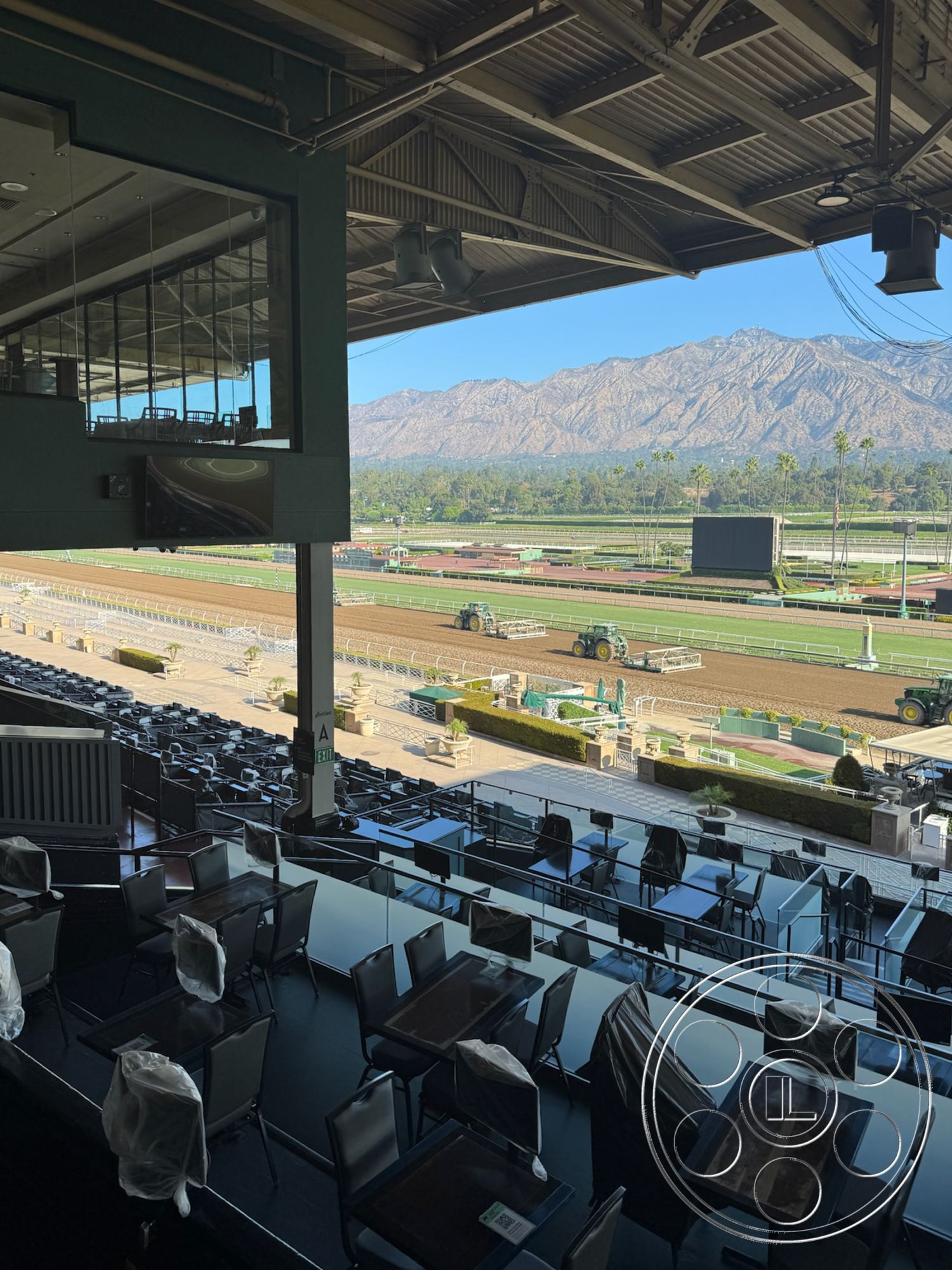 Arcadia Race Track - concrete flooring, exterior seating area, industrial architectural style, mountain view backdrop, covered outdoor space, metal framed glass windows, steel structure with exposed beams, open-air viewing terrace, modern racecourse facility, table seating arrangement, lush landscaping with shrubbery, urban horse racing venue, commercial grade furniture, panoramic viewing area, black powder-coated steel railings