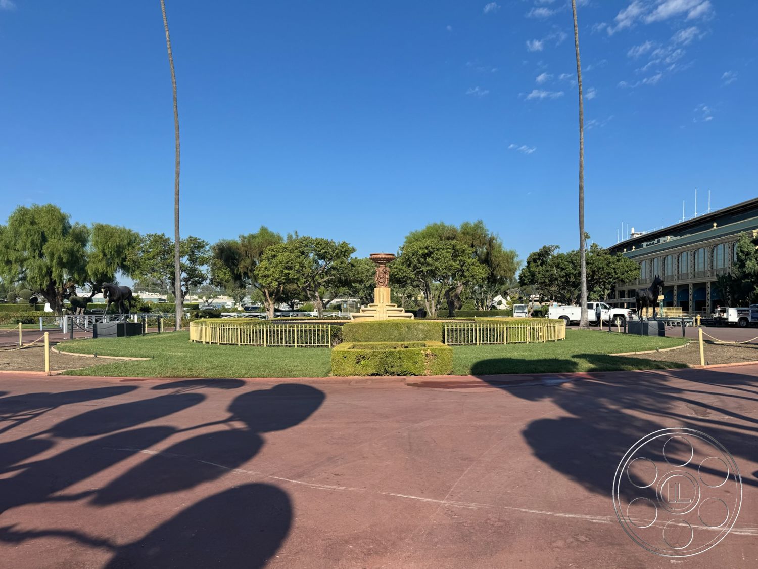 Arcadia Race Track - palm trees, outdoor seating area, circular driveway, clear blue sky, symmetrical layout, well-maintained greenery, exterior landscape design, modern building facade, urban park setting, granite fountain base, lawn with ornamental hedges, paved red concrete surface, sculptural horse figures, ornamental iron fencing, surrounding green gardens