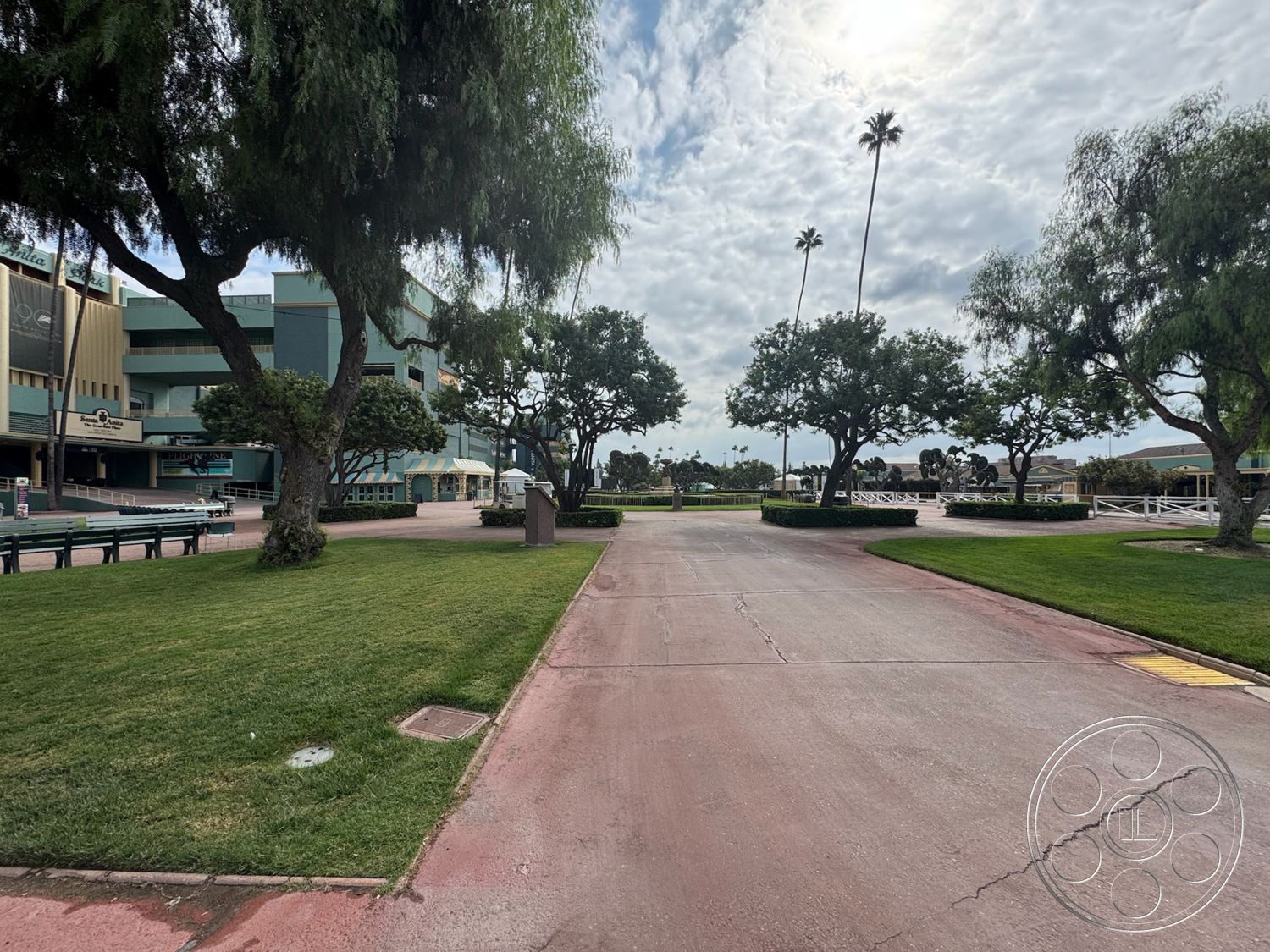 Arcadia Race Track - outdoor seating area, concrete walkway, symmetrical pathways, cloudy sky backdrop, open public space, commercial building exterior, urban park setting, palm tree lining, concrete landscaping curb, landscaped lawn with grass, organized exterior pathways, exterior building with large windows, decorative landscaping trees, well-maintained green areas