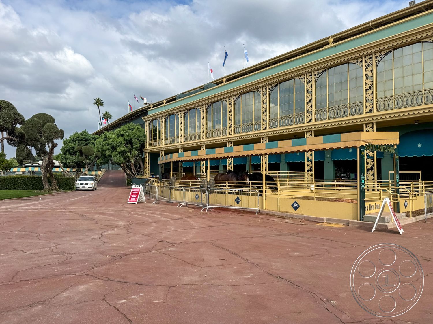 Arcadia Race Track - exterior, patio seating area, cultural landmark, vintage signage, colored concrete driveway, decorative wrought iron railings, large glass window panels, ornate architectural detailing, stylized concrete surface, historic racetrack building, garden setting with manicured trees, associated with horse racing, paved entrance walkway, vibrantly painted facade, multiple flags, southern California environment, sun-drenched atmosphere, open-air viewing area