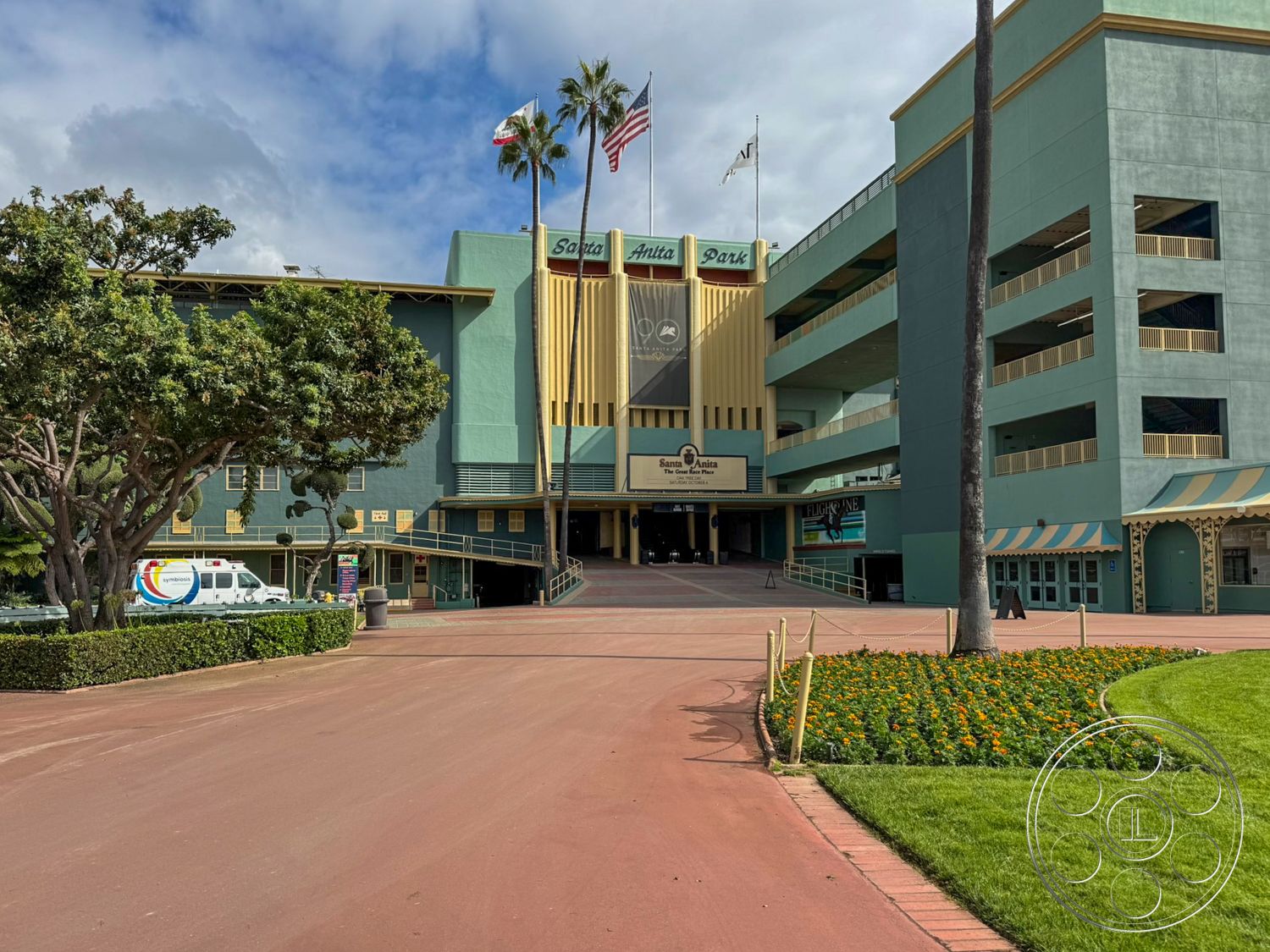 Arcadia Race Track - exterior, landscaped gardens, urban setting, paved walkway, modern architectural style, arched entryway, flag poles, flower bed, palm trees landscaping, concrete parking structure, painted concrete facade, green exterior wall, yellow accent details, parking lot entry, public park environment