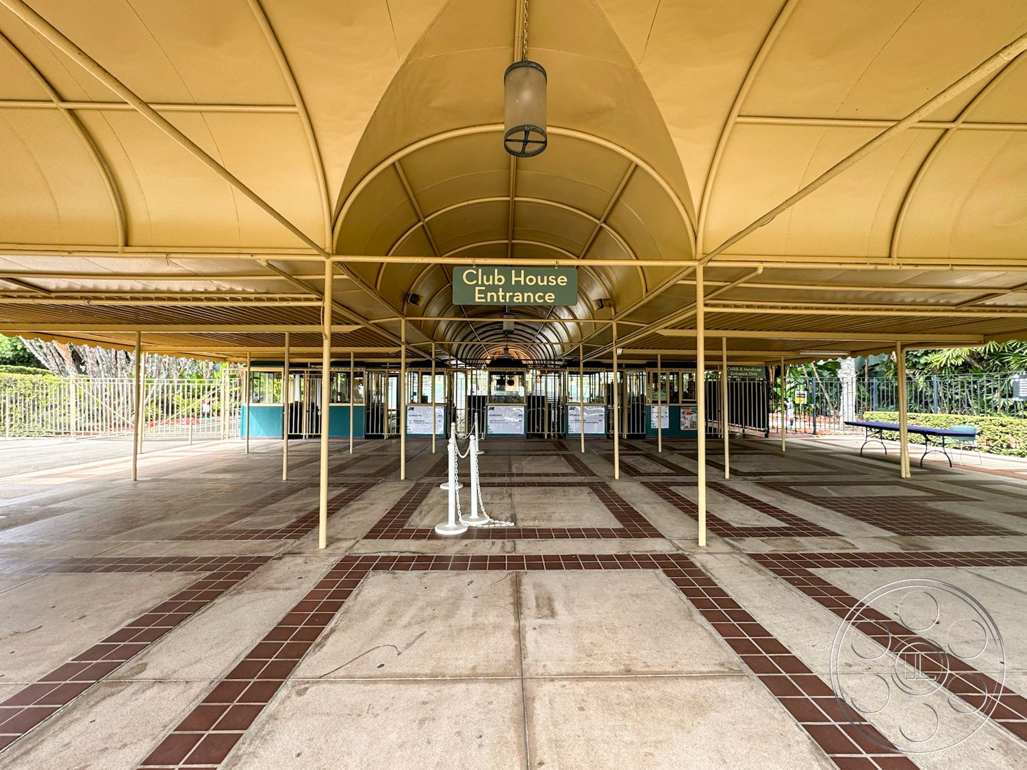 Arcadia Race Track - open space layout, metal railings, fenced perimeter, pedestrian-friendly design, open-air gathering space, exterior covered entrance, curved metal roof, concrete patio floor with tile inlays, red clay tile accents, bricked walkway, signage with green background, tropical landscaped surroundings, clubhouse entrance area, lamps suspended from ceiling supports, potted plants near entrance