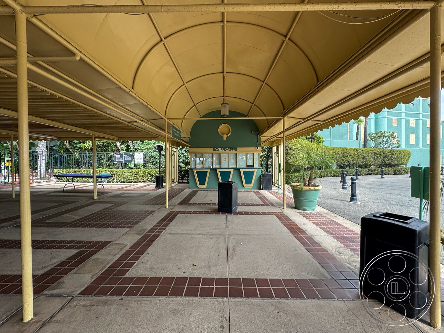 Arcadia Race Track - exterior, outdoor seating area, open-air design, accessible layout, lush greenery backdrop, concrete floor surface, decorative light poles, surrounding hedges, curved canopy structure, yellow fabric awning, brick-patterned pavers, ticket booth with teal exterior, tropical landscape with palm accents, trash bin for waste management, public gathering space, transitional space entry