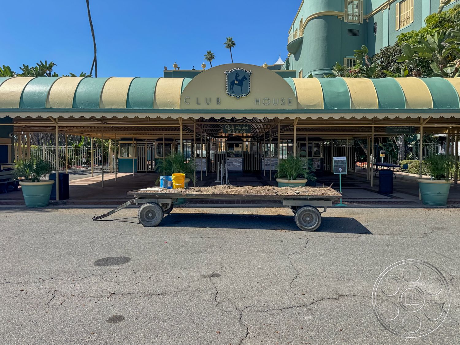 Arcadia Race Track - exterior, outdoor seating area, landscaped garden area, ground-level access, metal gate entrance, brick walkway leading to entrance, green and yellow striped awning, large planter pots with tropical plants, textured concrete pavement, clubhouse building with historic architecture, palm trees in surrounding landscape, signage with 'Club House' title