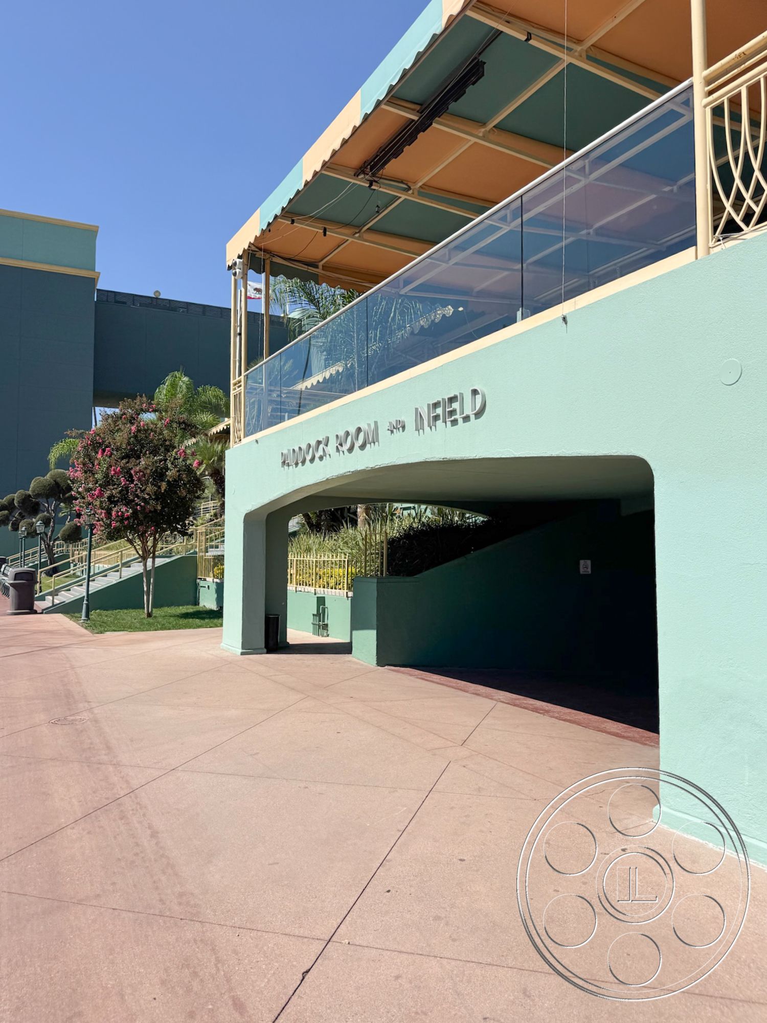 Arcadia Race Track - smooth concrete surface, public gathering area, open air space, accessible entryway, contemporary architectural style, shaded patio area, exterior concrete walkway, glass balcony railing, outdoor seating space, paved entrance area, painted green exterior walls, surrounding landscaping with palm trees, multifunctional pavilion structure, bright sunlit scene, nearby flowering shrubs
