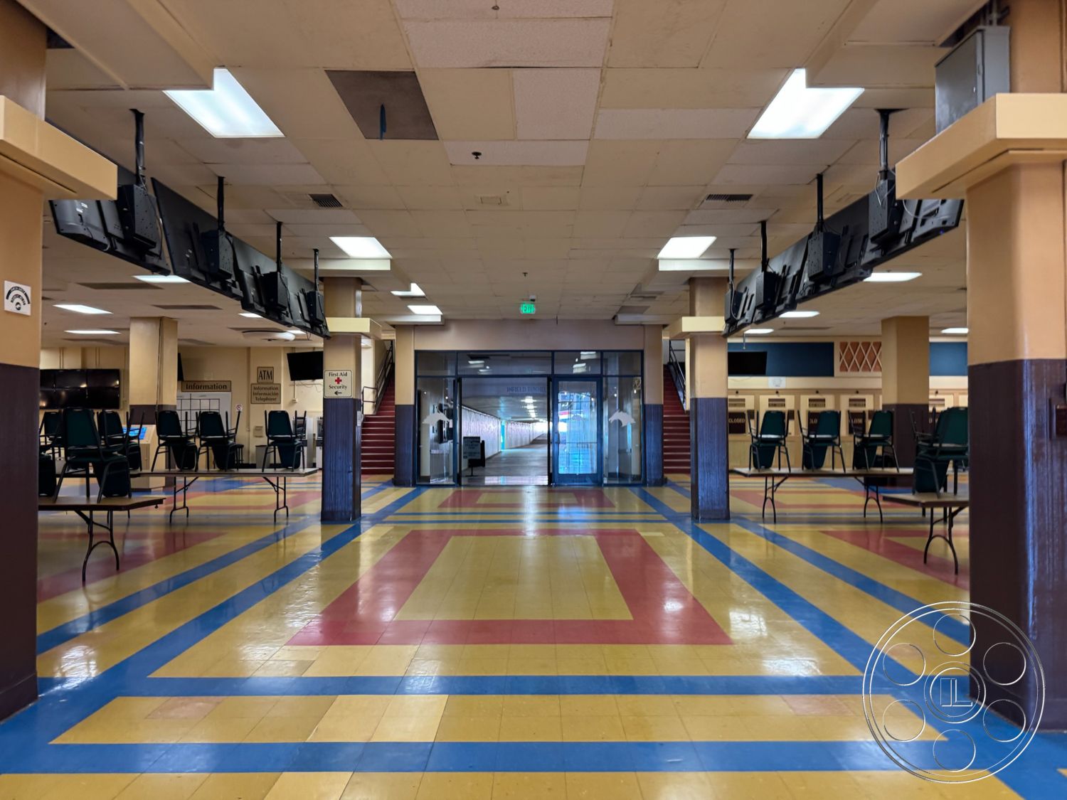 Arcadia Race Track - large windows for natural light, neutral wall paint, open floor plan layout, glass entry doors, commercial interior space, vinyl sheet flooring with colorful pattern, exposed ceiling tiles with fluorescent lighting, folding tables for seating arrangements, staircase with red carpet runner, temporary seating area with folding chairs, signage for information display, functional space for gatherings, public facility interior
