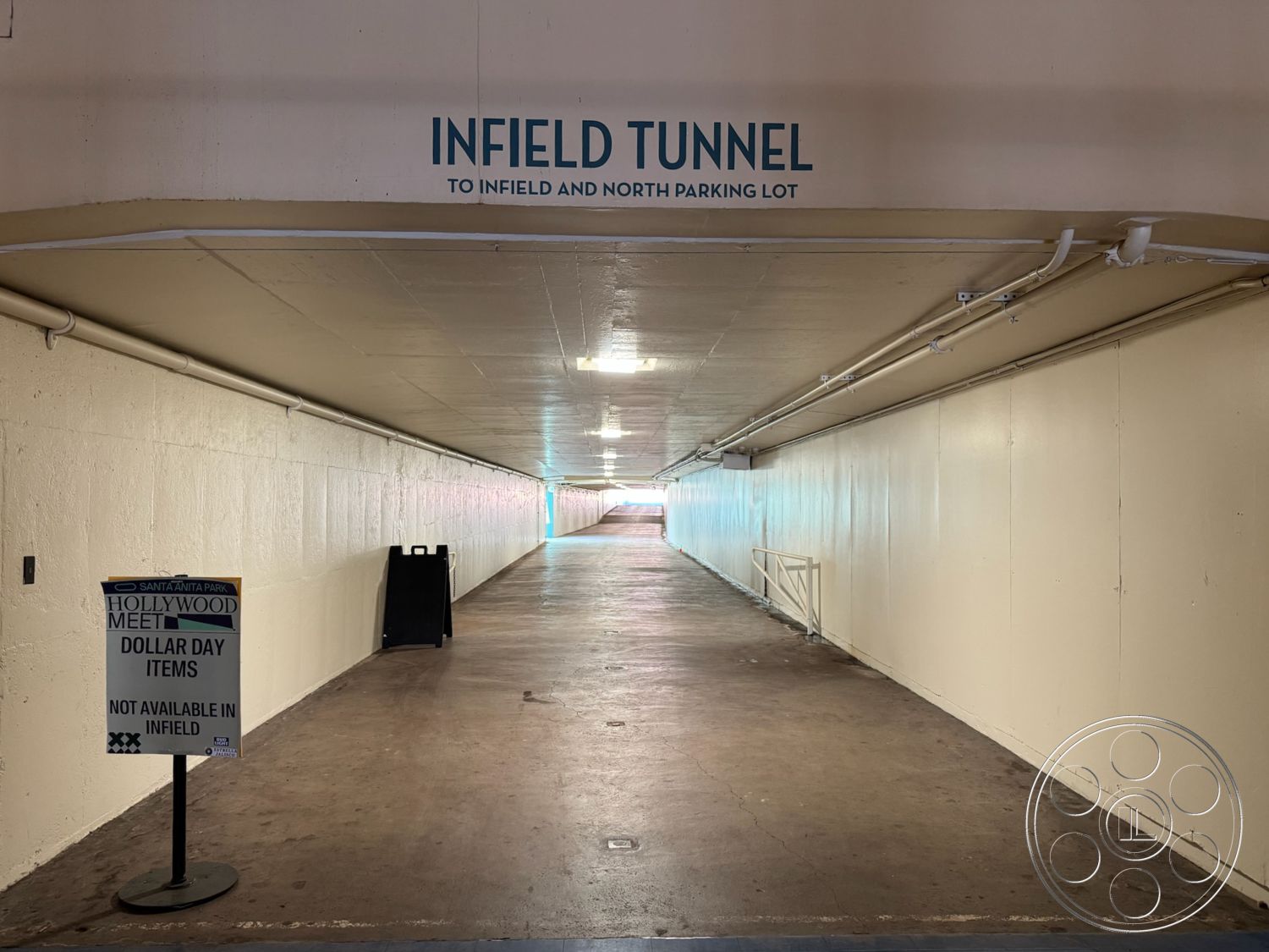 Arcadia Race Track - industrial lighting fixtures, modern minimalistic design, fluorescent ceiling lights, painted concrete walls, exposed concrete tunnel floor, rectangular tunnel shape, metal railing along tunnel, signage for parking lot, interior tunnel space, plain ceiling finish, level surface pathway