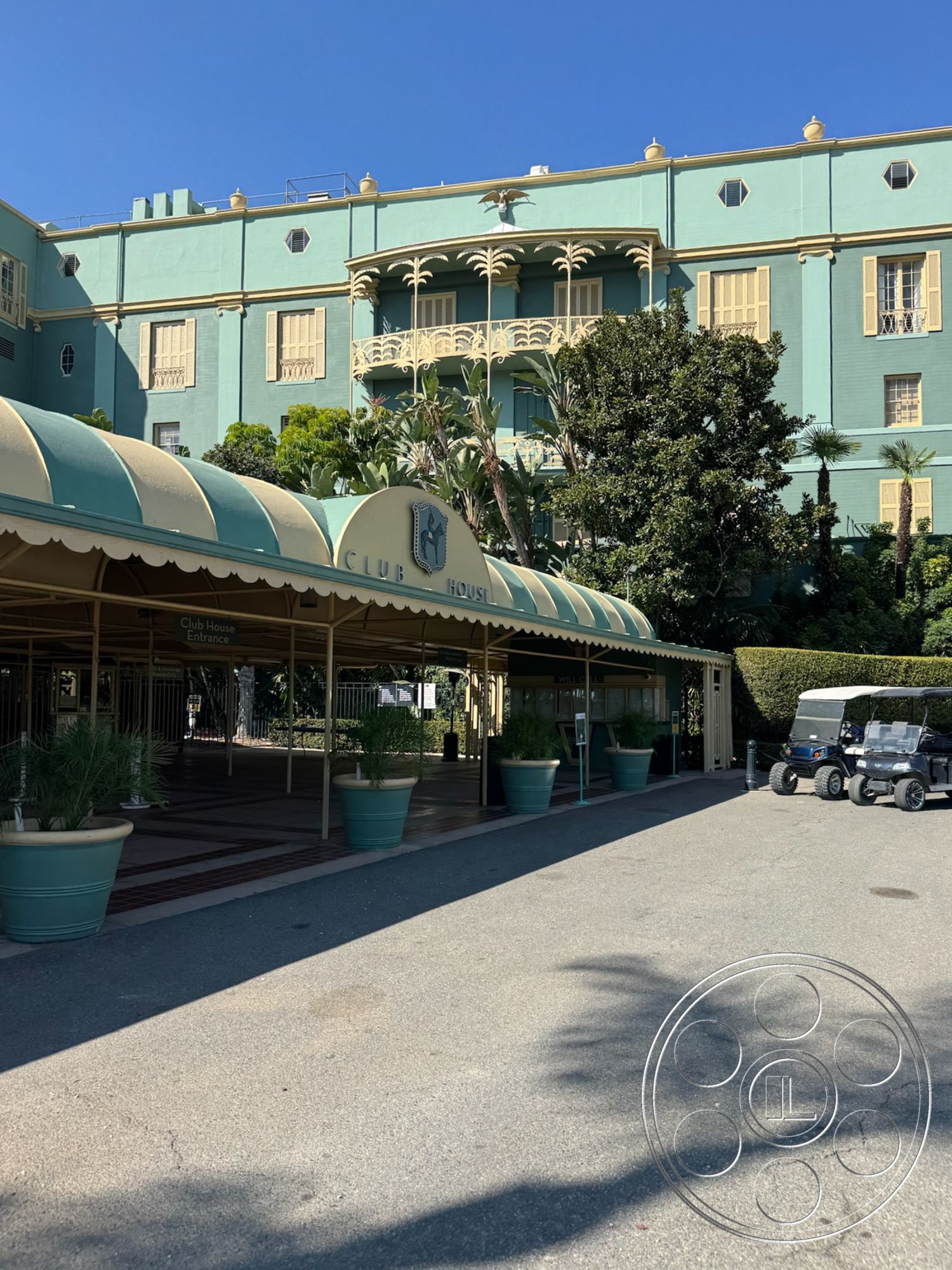 Arcadia Race Track - exterior view, stucco facade, red brick pathway, large potted plants, double-hung windows, decorative wrought iron railing, palm tree landscaping, courtyard style layout, symmetrical building design, arched entrance awning, clubhouse architectural detail, blue exterior paint, porch with canopy, golf cart parking area, sunny resort setting, vintage style architecture, shadow play from palm trees, perimeter fencing, outdoor entrance area