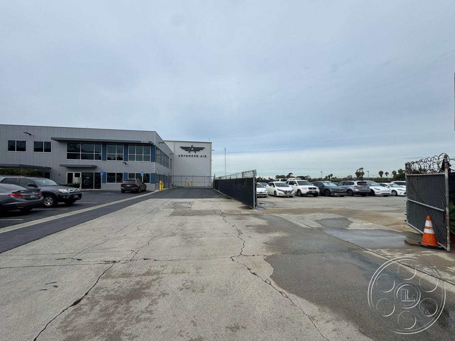 HW Hangar - exterior, large glass windows, urban setting, commercial building, asphalt driveway, functional architecture, metal facade, concrete parking lot, flat roof structure, industrial-style building, gated entrance with barbed wire, paved vehicle access, surrounding asphalt area, corporate office space