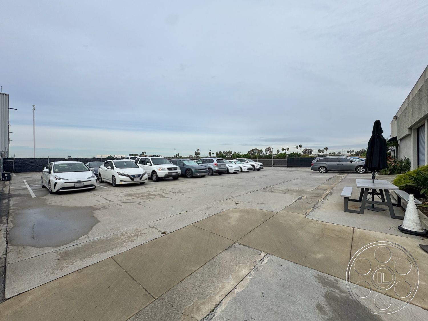 HW Hangar - exterior, urban parking lot, concrete parking lot surface, commercial building facade, palm tree surroundings, gray concrete pavers, city skyline view in distance, curved concrete driveways, hardsurface vehicle parking area, black metal fencing, landscaped patio area with umbrella, outdoor picnic table