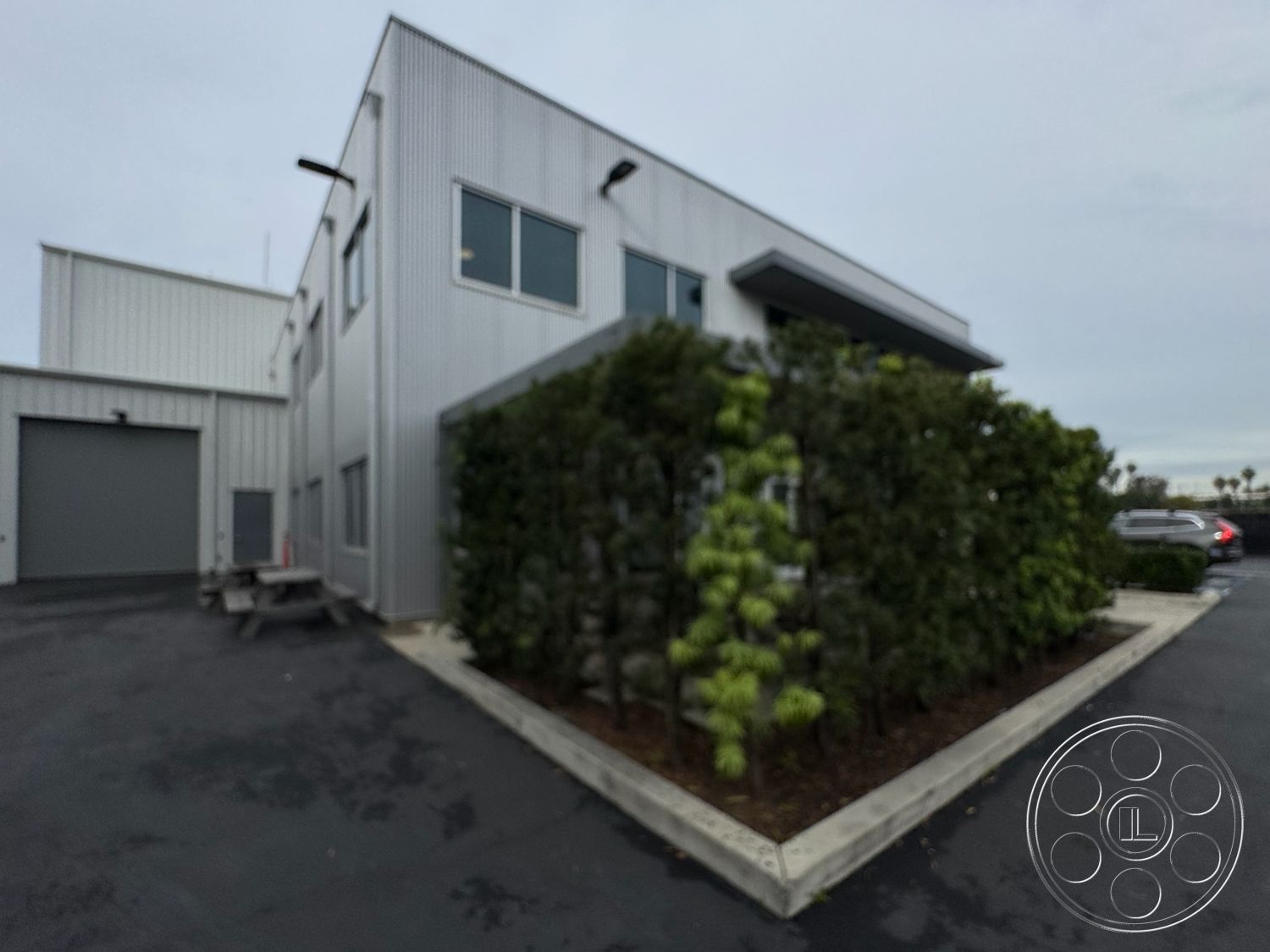 HW Hangar - flat roof design, large rectangular windows, minimalist architectural style, integrated outdoor seating, surrounding urban environment, commercial property setting, modern industrial architecture, landscaped garden border, exterior steel panel siding, concrete planter beds, asphalt surface parking area, secluded courtyard area