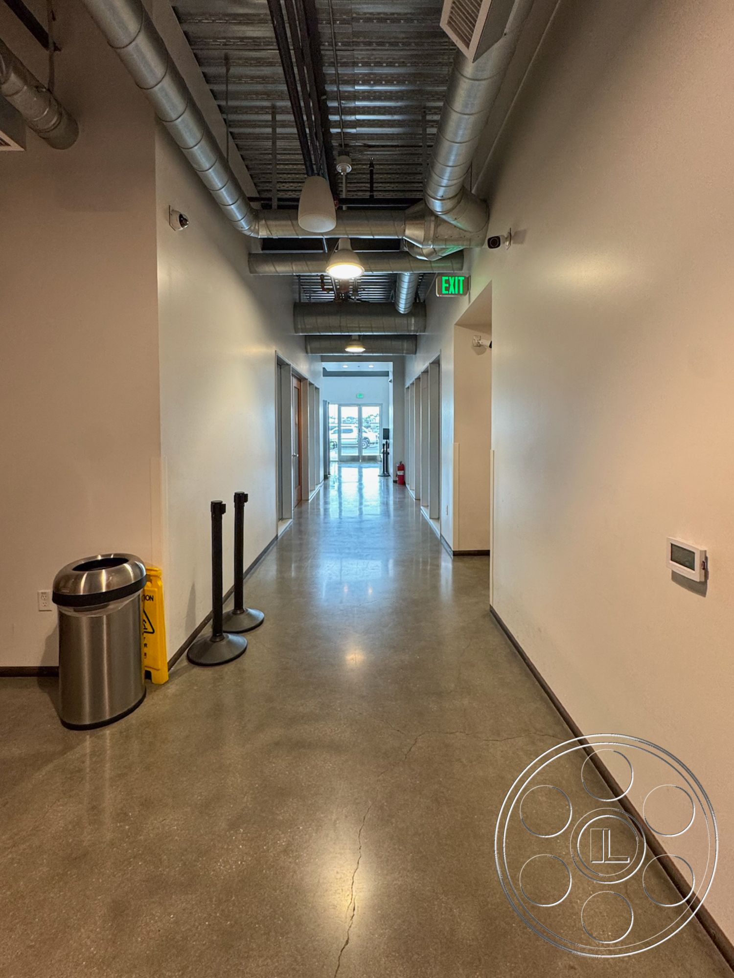 HW Hangar - exposed steel beams, acoustic ceiling tiles, neutral painted walls, industrial style interior, contemporary office environment, metal ceiling ductwork, minimalist interior space, smooth concrete wall finish, clean lines architectural style, functional public space, commercial hallway design, fire safety equipment, polished concrete hallway floor, flush wall-mounted lighting fixtures, corridor with glass exit doors, modern trash receptacle, stainless steel stanchions, wide open entryway