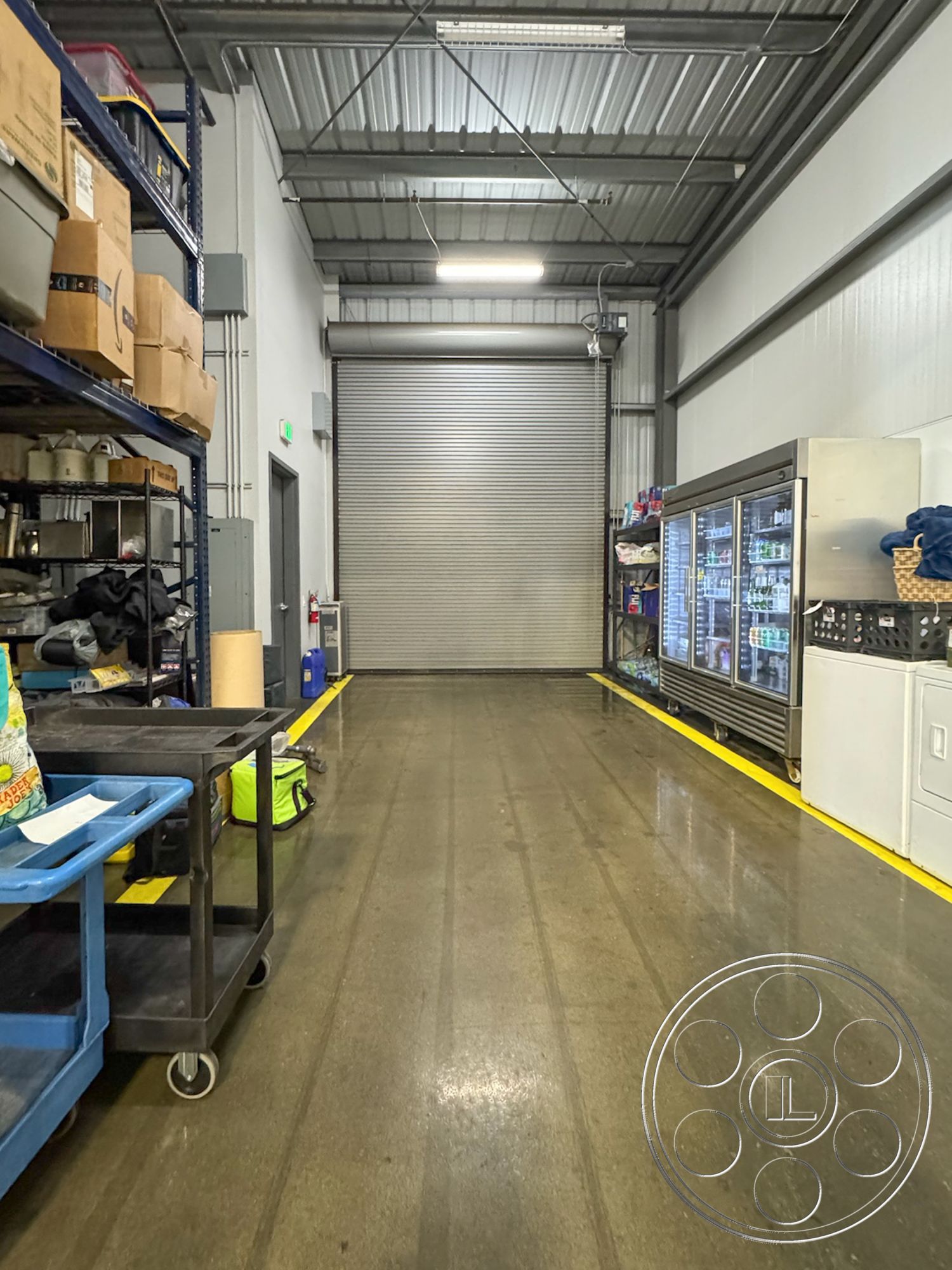 HW Hangar - stainless steel appliances, large open space, concrete polished floor, interior warehouse space, warehouse storage area, industrial shelving units, ambient warehouse lighting, fire extinguisher mounted on wall, refrigerated display case, storage boxes on shelves, metal corrugated ceiling panels, roller shutter door, open commercial layout, utility cart with blue frame, yellow strip floor marking, cleaning supplies in visible storage