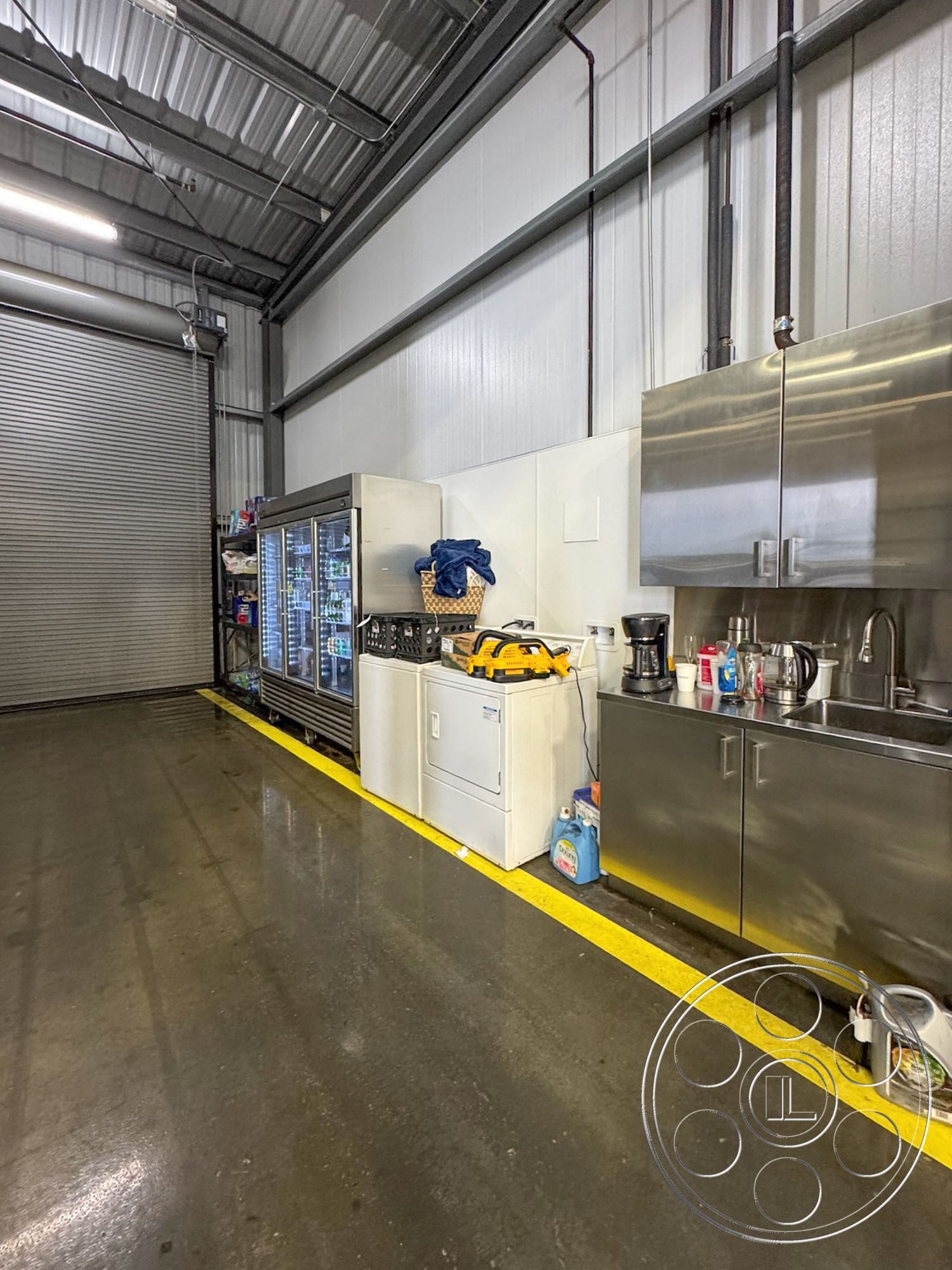HW Hangar - minimalist decor, interior kitchen space, stainless steel countertop, bright overhead lighting, functional utility space, concrete floor with epoxy finish, metal ceiling panels, industrial style cabinetry, refrigerated glass display case, white washing machine, yellow safety line on floor, plumbing pipes on wall, modern coffee maker, open storage shelves, industrial kitchen equipment