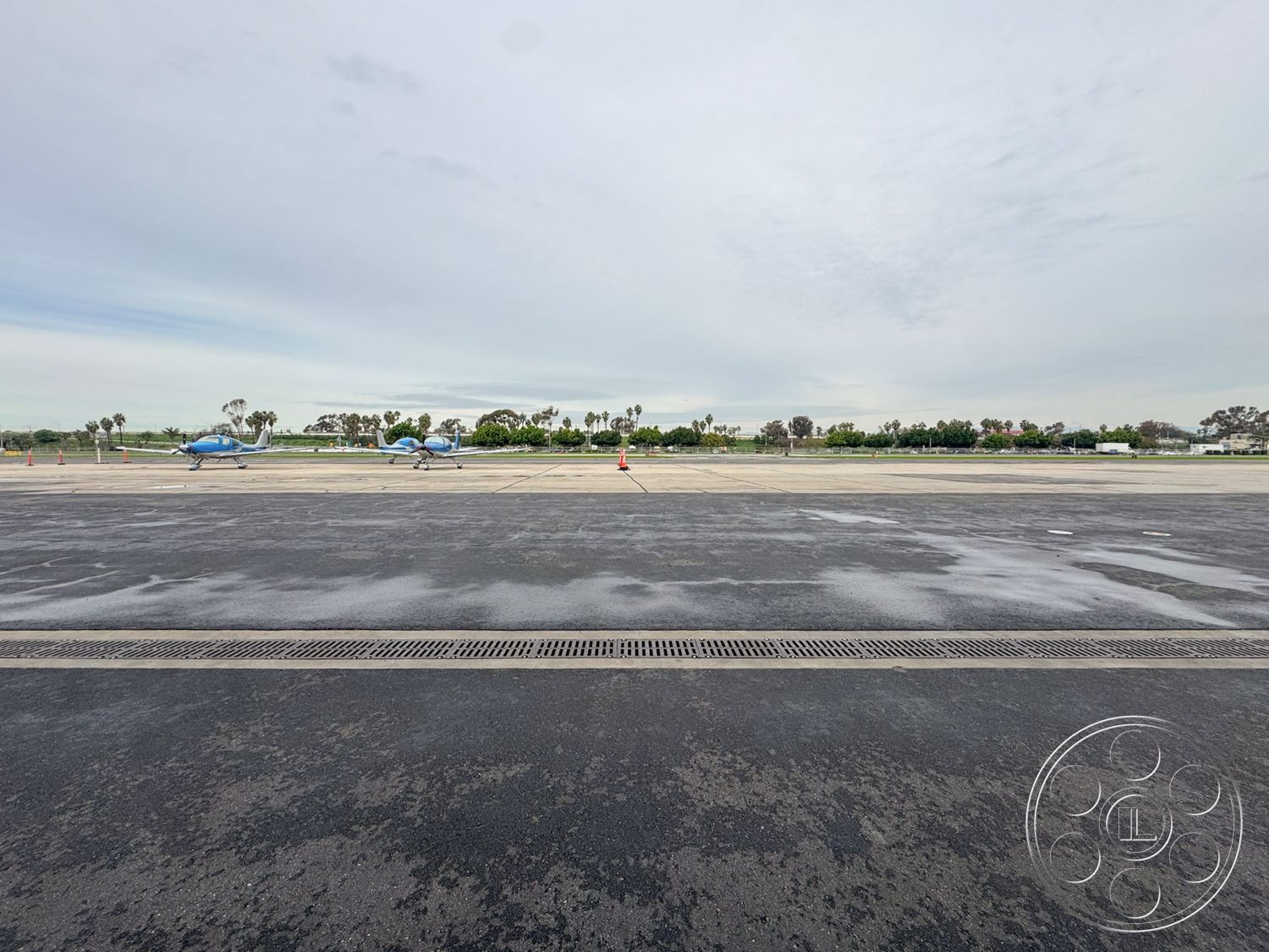 HW Hangar - palm tree landscaping, landscaped green spaces, asphalt runway surface, helicopter parking area, drainage grates on pavement, overcast sky exterior, airport tarmac setting