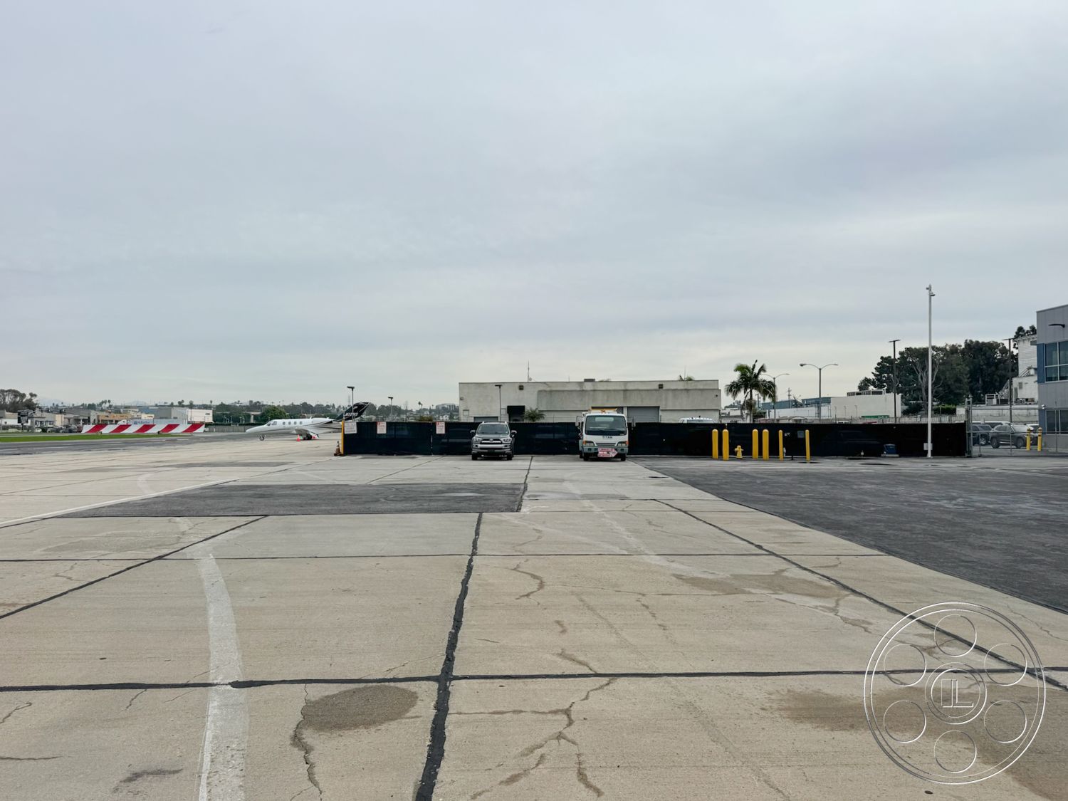 HW Hangar - exterior view, asphalt parking lot, exposed aggregate concrete, cloudy sky backdrop, urban airport setting, concrete airport runway, modern industrial building, metal fence perimeter, tarmac surface, rectangular parking spaces, curved drive lanes, steel light poles, paved taxiway, concrete loading docks, functional airport infrastructure, vehicle access area