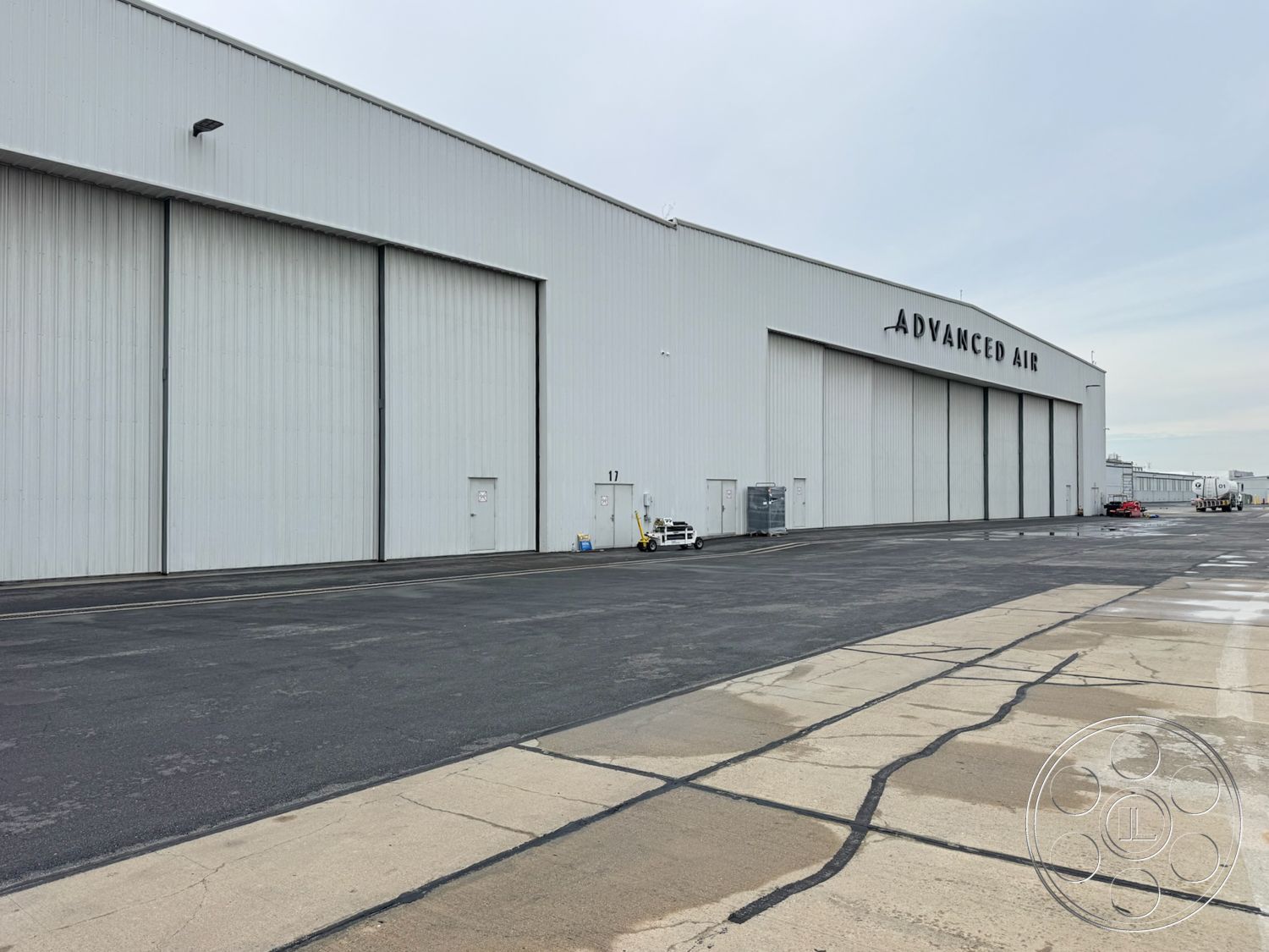 HW Hangar - overhead lighting fixtures, flat roof design, surrounding airport environment, large bi-fold hangar doors, exterior steel hangar structure, metal siding cladding, concrete apron for aircraft parking, industrial warehouse facade, white exterior paint finish, large exterior signage, asphalt surface for parking, drainage channels along pavement, access service doors, expansive open space layout