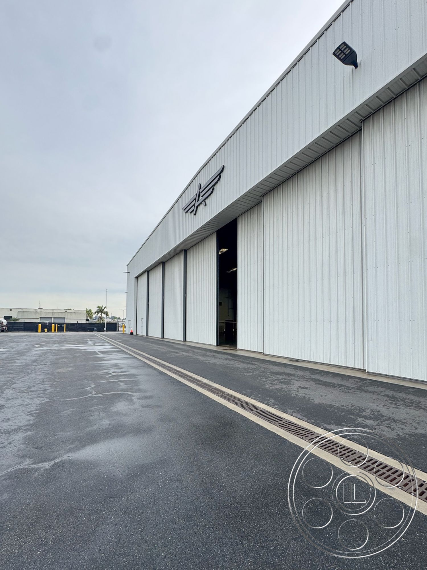 HW Hangar - industrial architecture, asphalt parking lot, functional outdoor area, exposed structural elements, flat roof design, steel frame construction, commercial building exterior, reinforced concrete foundation, minimalist facade, modern industrial aesthetic, large hangar doors, high bay doors, metal exterior cladding, airport hangar setting, urban airport environment, drainage trench, glossy concrete finish, wide entrance pathway, weathered exterior, open space for aircraft handling