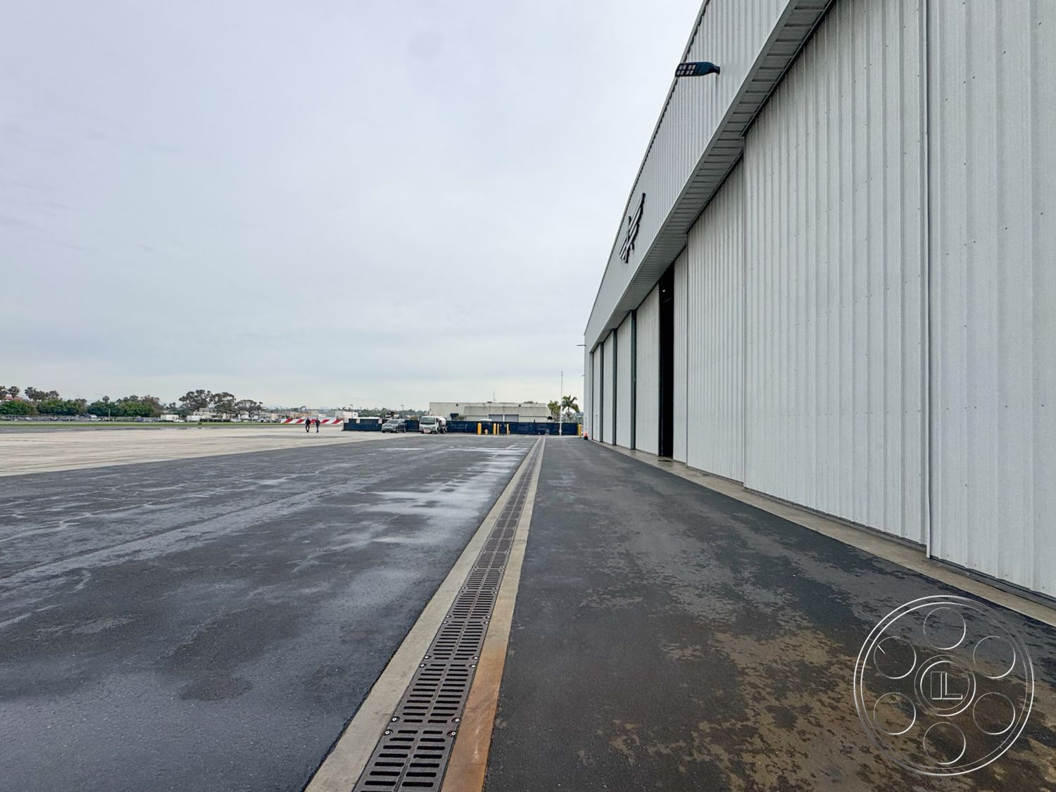 HW Hangar - corrugated metal siding, textured concrete surface, cloudy sky backdrop, urban industrial setting, surrounding airport environment, exterior concrete pavement, industrial steel hangar structure, asphalt apron area, drainage trench along driveway, large loading doors, service access pathway, flat-roofed architecture, open-air aviation area