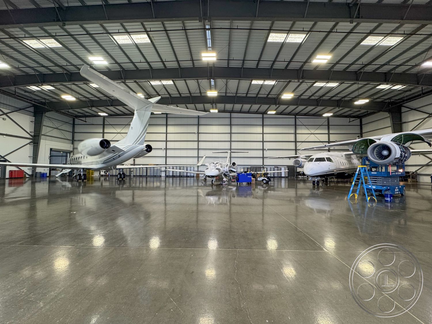 HW Hangar - bright fluorescent lighting, concrete polished floor, exposed steel structural elements, steel framed aircraft hangar interior, industrial style open layout, high vaulted ceiling with metal trusses, large hangar doors for aircraft access, hangar work area with tool carts, multiple private jets parked, efficient workspace design for aircraft maintenance, durable and functional hangar environment, spacious aircraft storage area