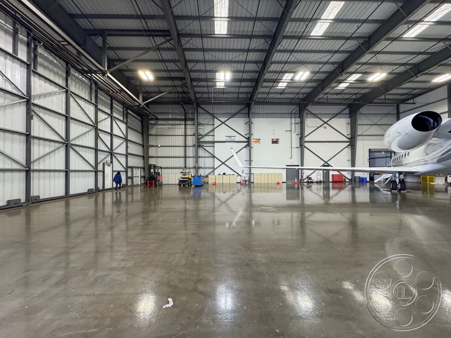 HW Hangar - high ceilings, open floor plan, polished concrete flooring, exposed metal beams, utility shelving, natural light from skylights, safety cones, functional industrial design, aircraft maintenance area, exterior steel structure, industrial hangar interior, large metal doors, forklift operational space, hangar space for private jet, white insulated metal wall panels, clean, spacious environment, aircraft storage and servicing area