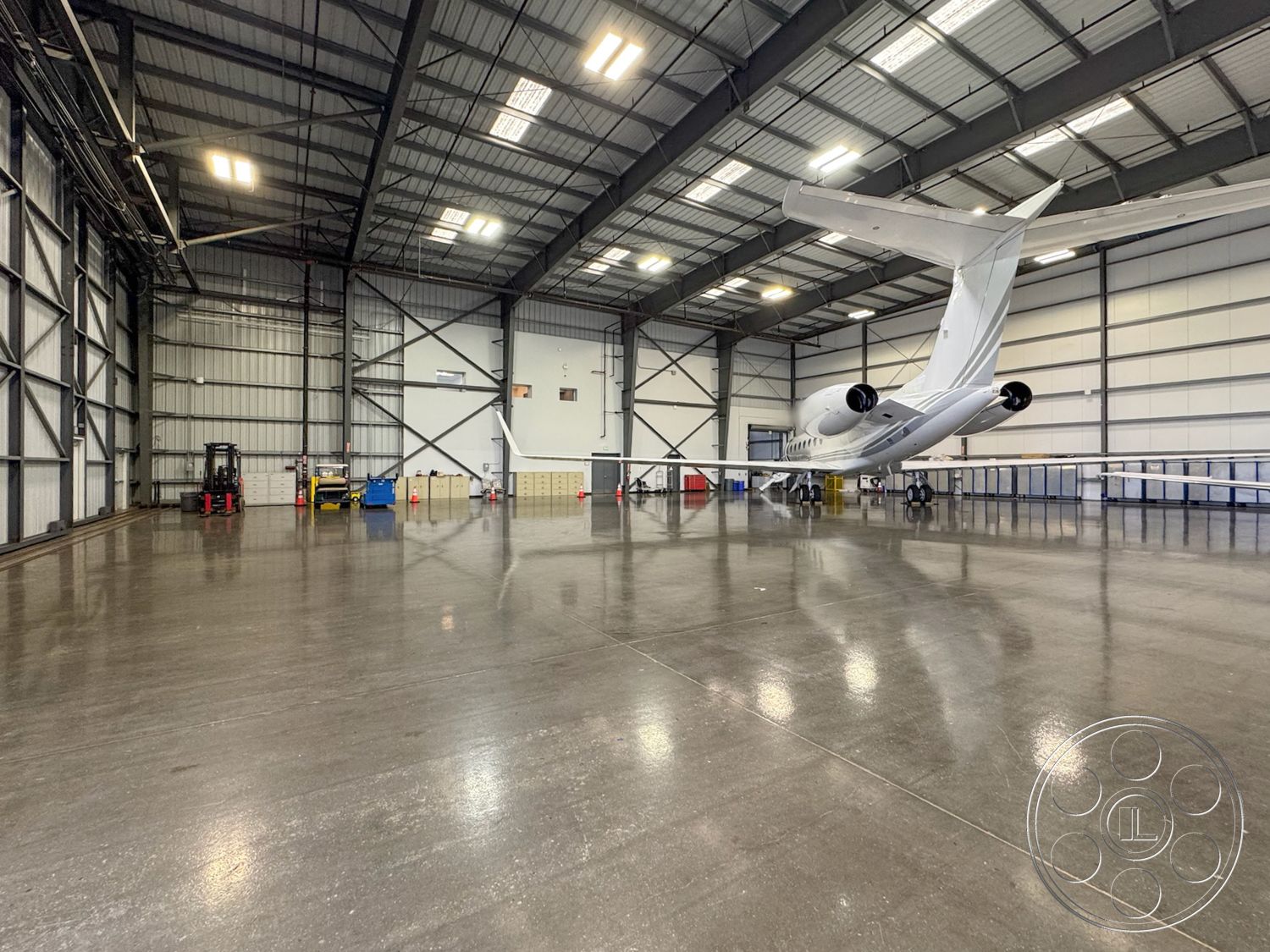 HW Hangar - polished concrete flooring, industrial lighting, exposed steel beams, concrete floor finish, metal roof structure, aircraft maintenance space, high ceiling industrial space, large sliding hangar doors, steel framework interior, hangar space with airplane, open layout aviation facility, catwalk lighting fixtures, service vehicle storage area, mechanical equipment storage, large open workspace, airplane parking space