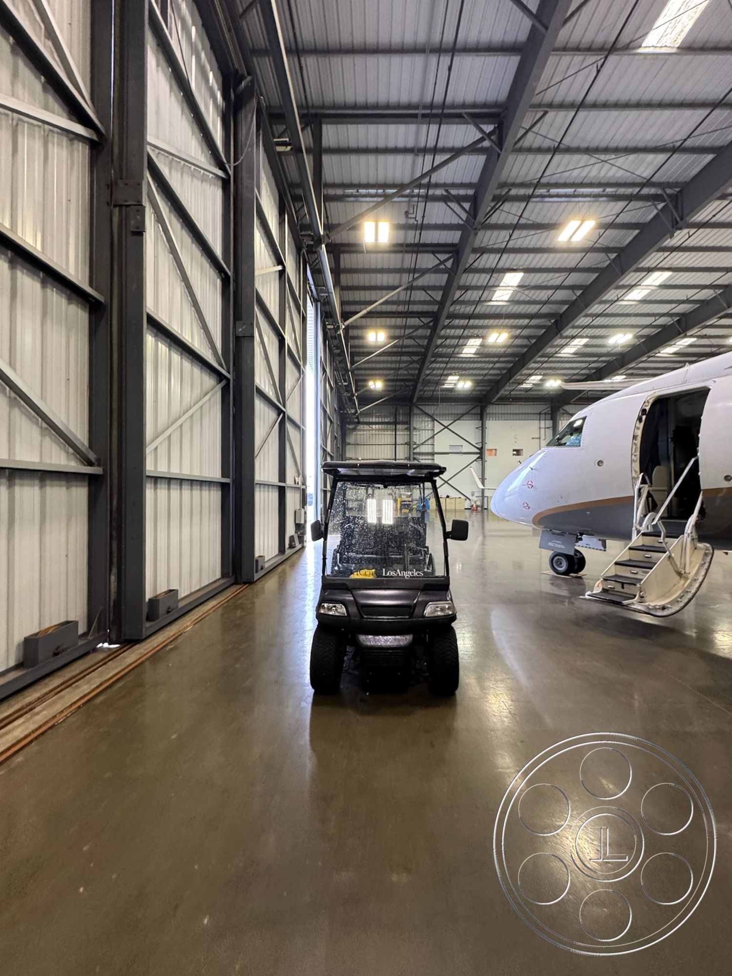 HW Hangar - industrial lighting fixtures, exposed steel beams, concrete wall finish, high ceiling with metal trusses, aircraft maintenance space, large hangar doors, metal frame structure, modern aviation facility, interior hangar space, golf cart parking area, commercial aviation environment, solid concrete flooring, wide open space interior, plane access area, functional layout design, spacious and utilitarian, high-traffic operational zone