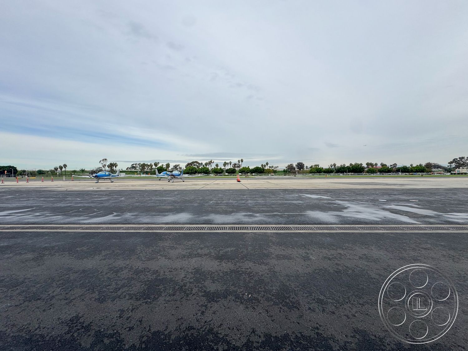 HW Hangar - surrounding palm trees, asphalt runway surface, concrete drainage grates, tarmac airport apron, paved helipad area, landscaped airport environment, outdoor aviation area, aircraft parking zone, curved taxiway design, overcast sky setting, urban airport location, exterior airport facilities, view of aircraft hangars, multi-plane layout, open air aviation space