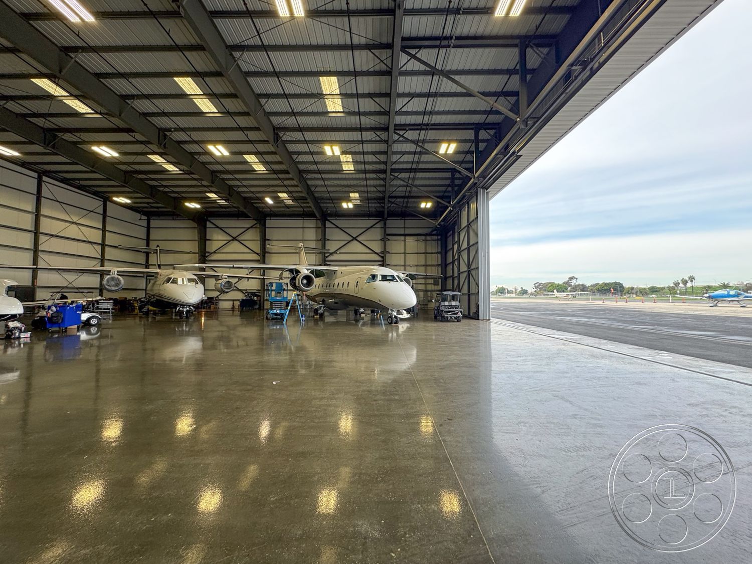 HW Hangar - industrial lighting fixtures, open floor layout, concrete flooring with polished finish, exterior hangar structure, aircraft parking area, urban airport setting, maintenance equipment storage, large sliding hangar doors, aircraft servicing area, glass windows providing natural light, metal framing construction, high ceilings with exposed steel beams, working aircraft maintenance space, ground equipment storage, hangar office space, aircraft access pathway, nearby runway view