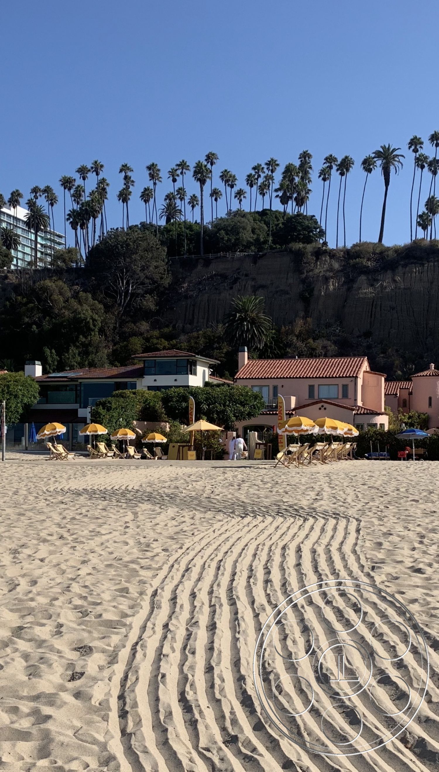 The Pink Beach House - exterior, terracotta roof tiles, beachfront property, palm tree landscaping, open-air dining area, sand beach floor, resort-style outdoor seating, yellow umbrella shading, colorful stucco walls, coastal architectural style, shoreline view, lush greenery around buildings, patio furniture with chaise lounges, textured sand pathways, sunlit beach setting, urban hillside background