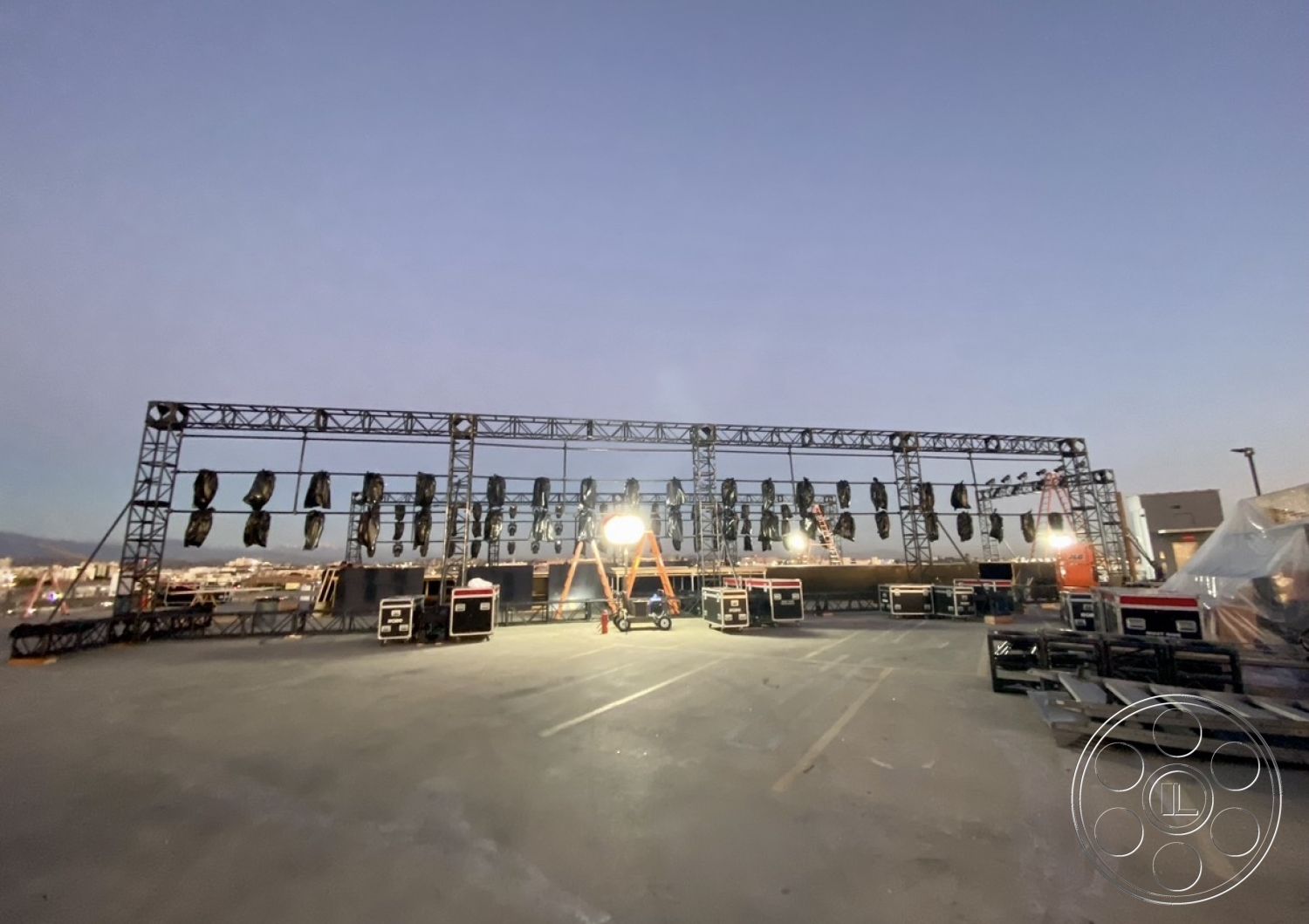 SKY Rooftop Garage - concrete parking surface, sound equipment storage, metal ladder, exterior stage structure, steel truss framework, LED stage lighting, outdoor performance area, industrial lighting rigging, temporary event setup, view of urban skyline at dusk, lighting equipment setup, event production area, stage backdrop, illuminated performance stage