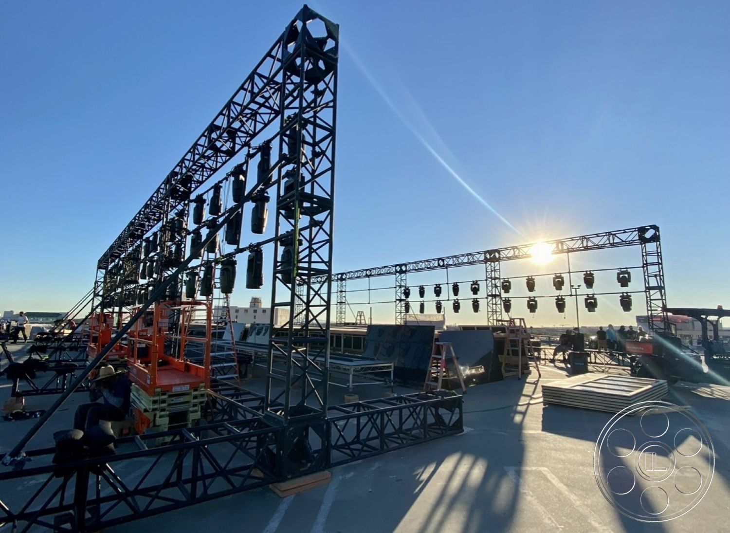 SKY Rooftop Garage - sunset backdrop, urban skyline view, steel truss framework, metal scaffolding structure, LED stage lights, aerial stage setup, tools and equipment for performance installation, open rooftop space, concert event preparation, industrial construction materials, elevated performance stage, exterior setup, lighting rig assembly, performance event staging, temporary installation architecture