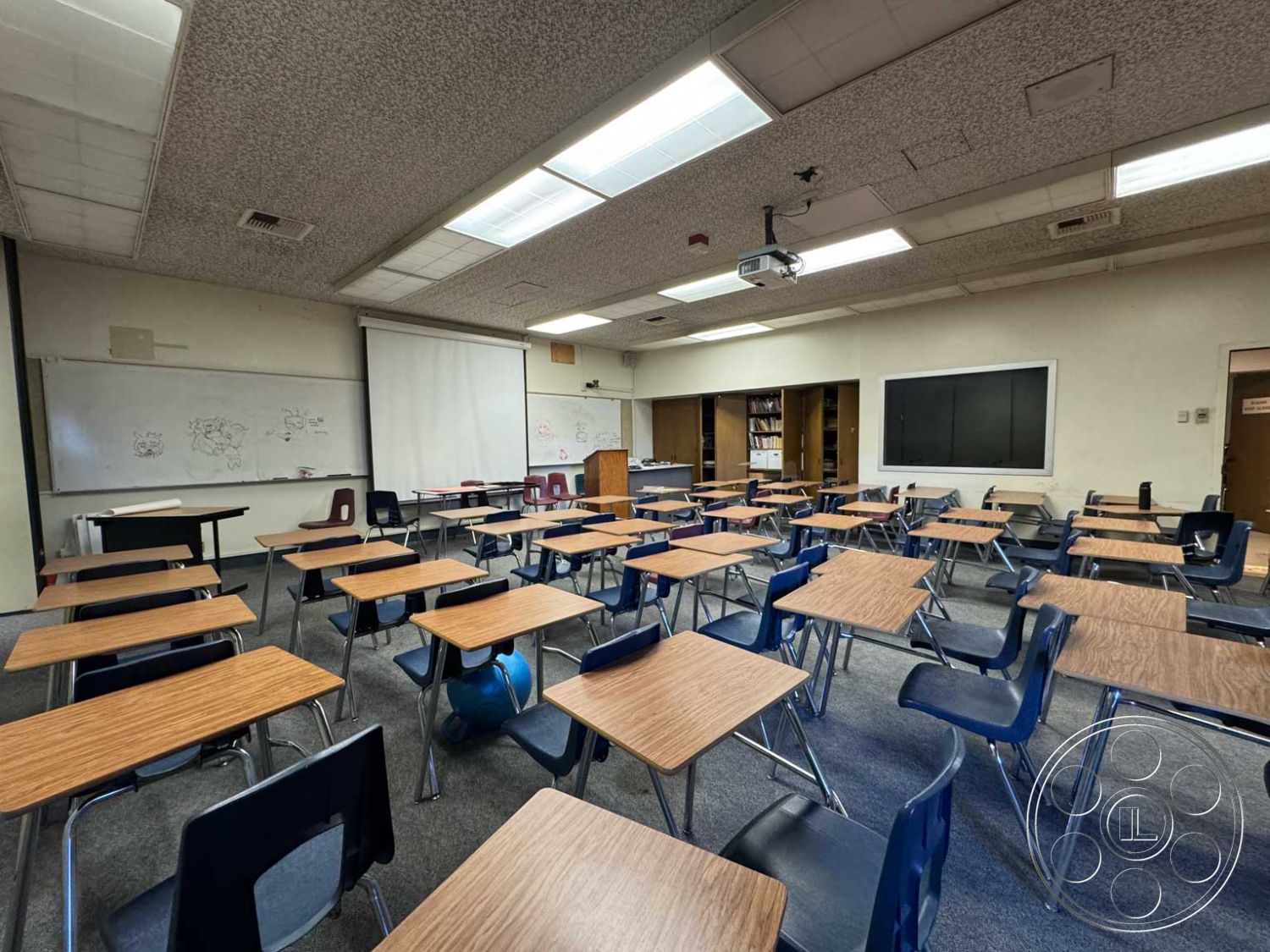 Old School - carpet flooring, projector screen, overhead fluorescent lighting, acoustic ceiling panels, painted drywall walls, interior classroom space, metal-framed student chairs, floating ceiling tiles, laminated wood classroom desks, interactive whiteboard wall, shelving unit for classroom materials, blackboard with white chalk markings, urban educational setting