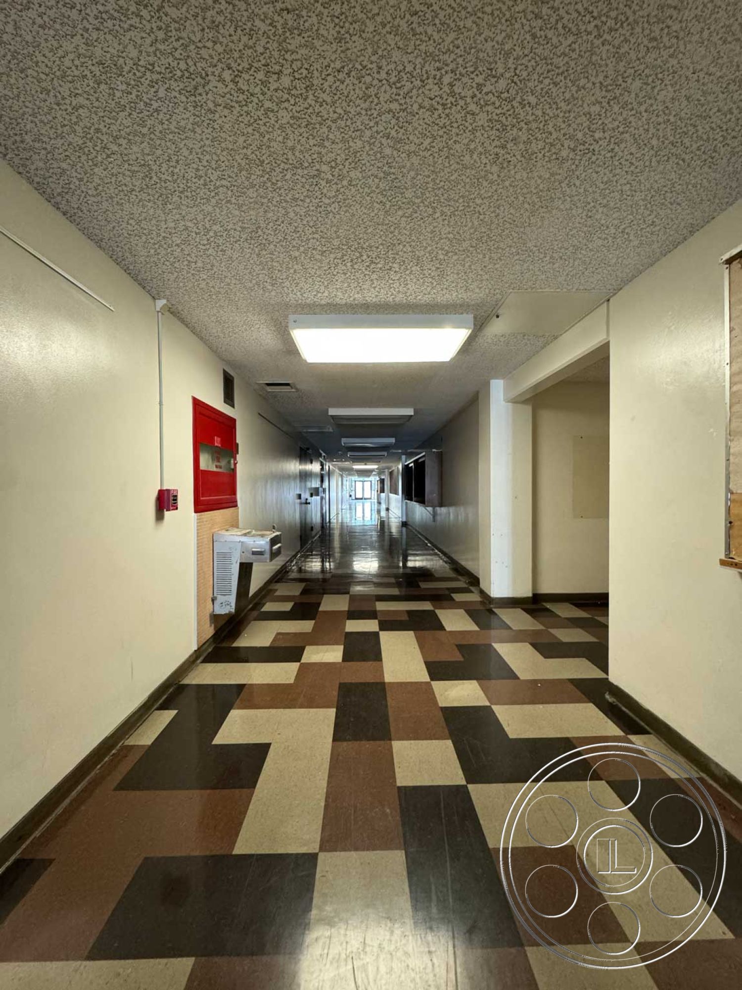 Old School - interior hallway, painted drywall walls, commercial building interior, polished vinyl flooring, fluorescent ceiling lights, textured ceiling tiles, industrial-style lighting, emergency exit signage, checkerboard pattern floor tiles, loading dock area, brown and beige color scheme, long corridor design, central heating unit, school building hallway, adjacent rooms access, functional architectural design
