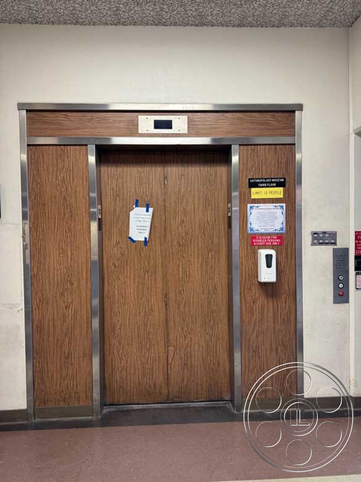 Old School - concrete floor, textured ceiling tiles, elevator control panel, exterior elevator entrance, wood panel elevator doors, stainless steel trim, yellow warning sign, ground floor location, public building accessibility, signage indicating limited capacity, elevator call button panel, informational notice on door, entrance to museum, ADA compliant features