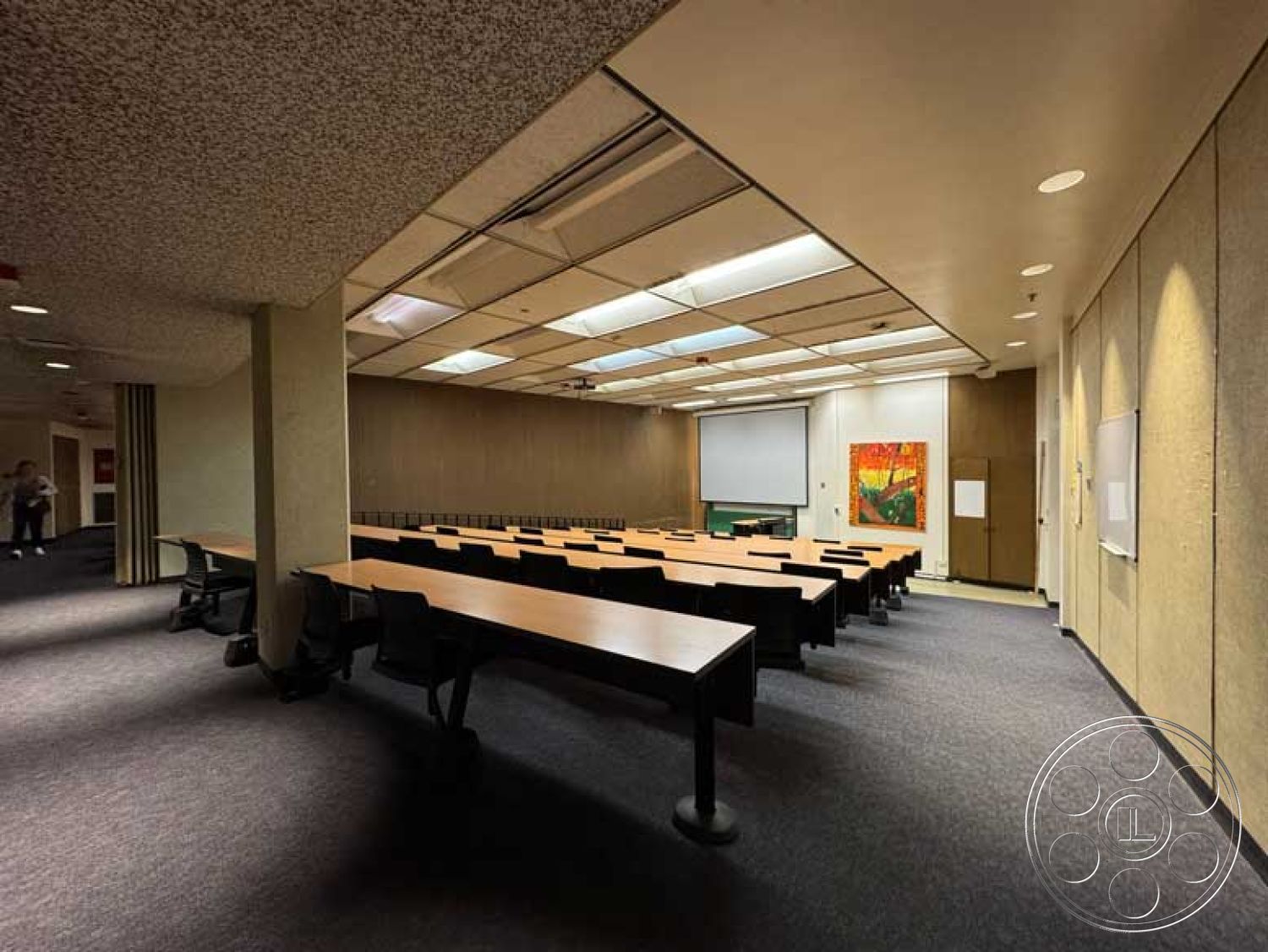 Old School - artwork on wall, carpeted flooring, textured plaster walls, dark wood paneling, black ergonomic chairs, acoustic ceiling panels, LED ceiling lights, large windows with blinds, interior classroom space, projector screen wall, rectangular wooden tables, dual access points