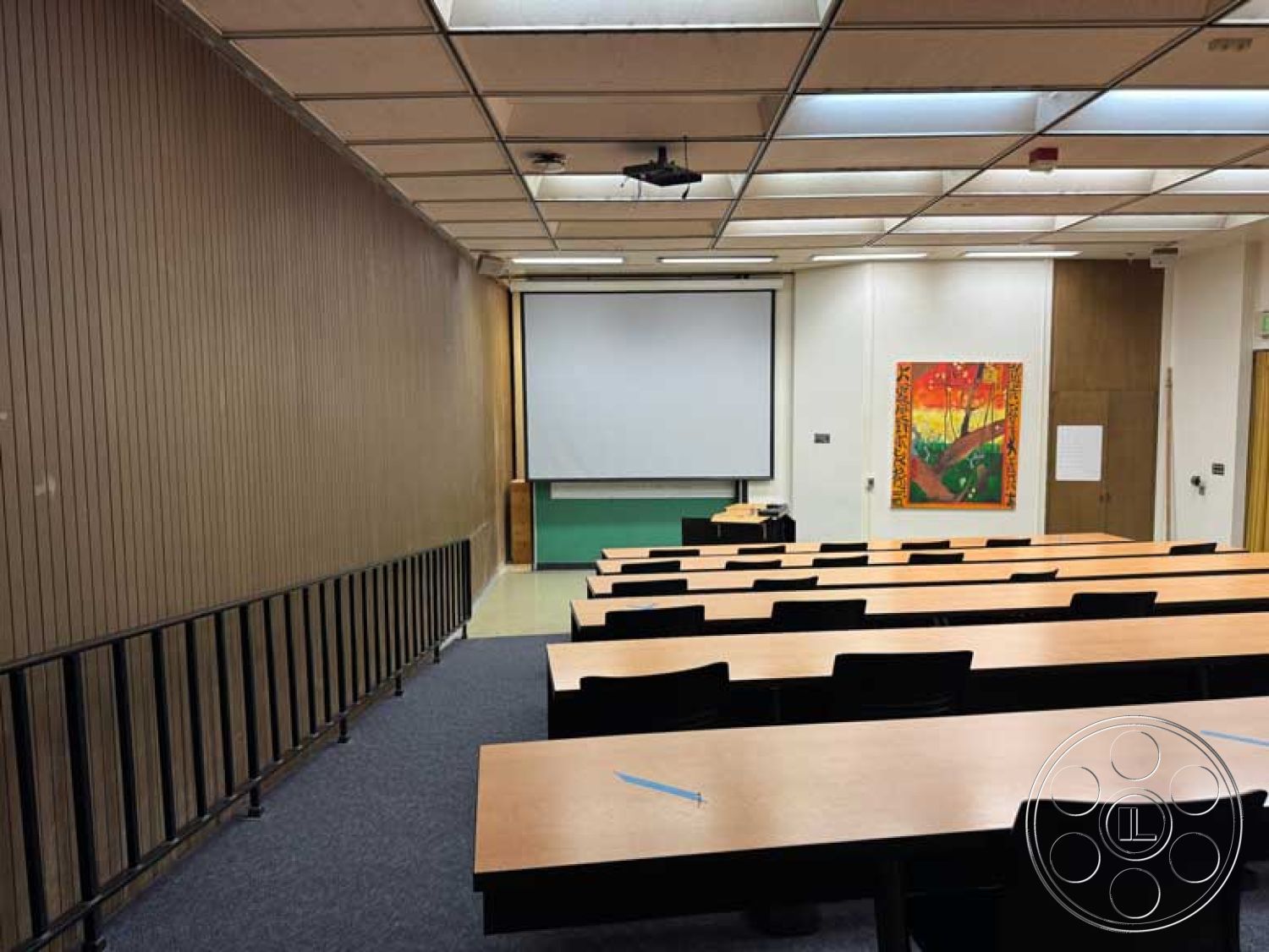 Old School - carpet flooring, acoustic ceiling tiles, wood paneling wall treatment, steel railing details, interior classroom design, wooden lecture tables, retractable projection screen, black molded plastic chairs, flat ceiling with fluorescent lighting, colorful wall artwork, chalkboard wall section, sightline-focused seating arrangement