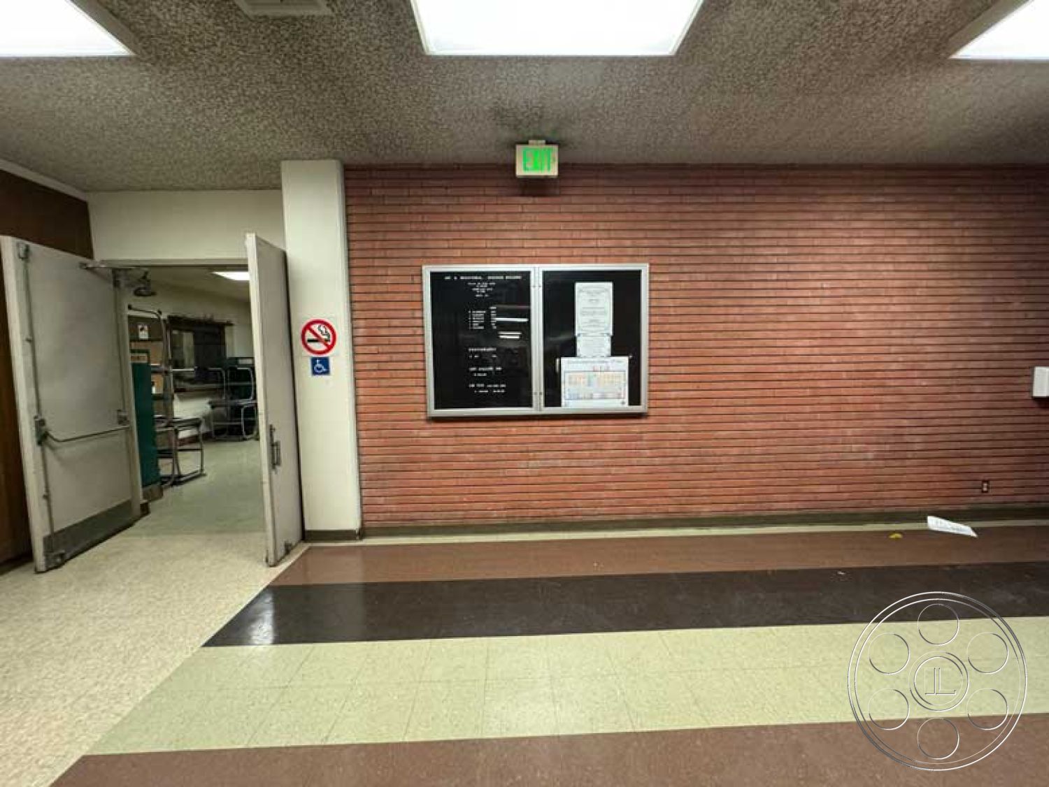 Old School - double door entry, acoustic ceiling tiles, exposed brick accent wall, fluorescent ceiling lights, commercial hallway interior, vinyl flooring tiles, black notice board with white paper, detection exit sign, green emergency exit light, curved tile flooring transition