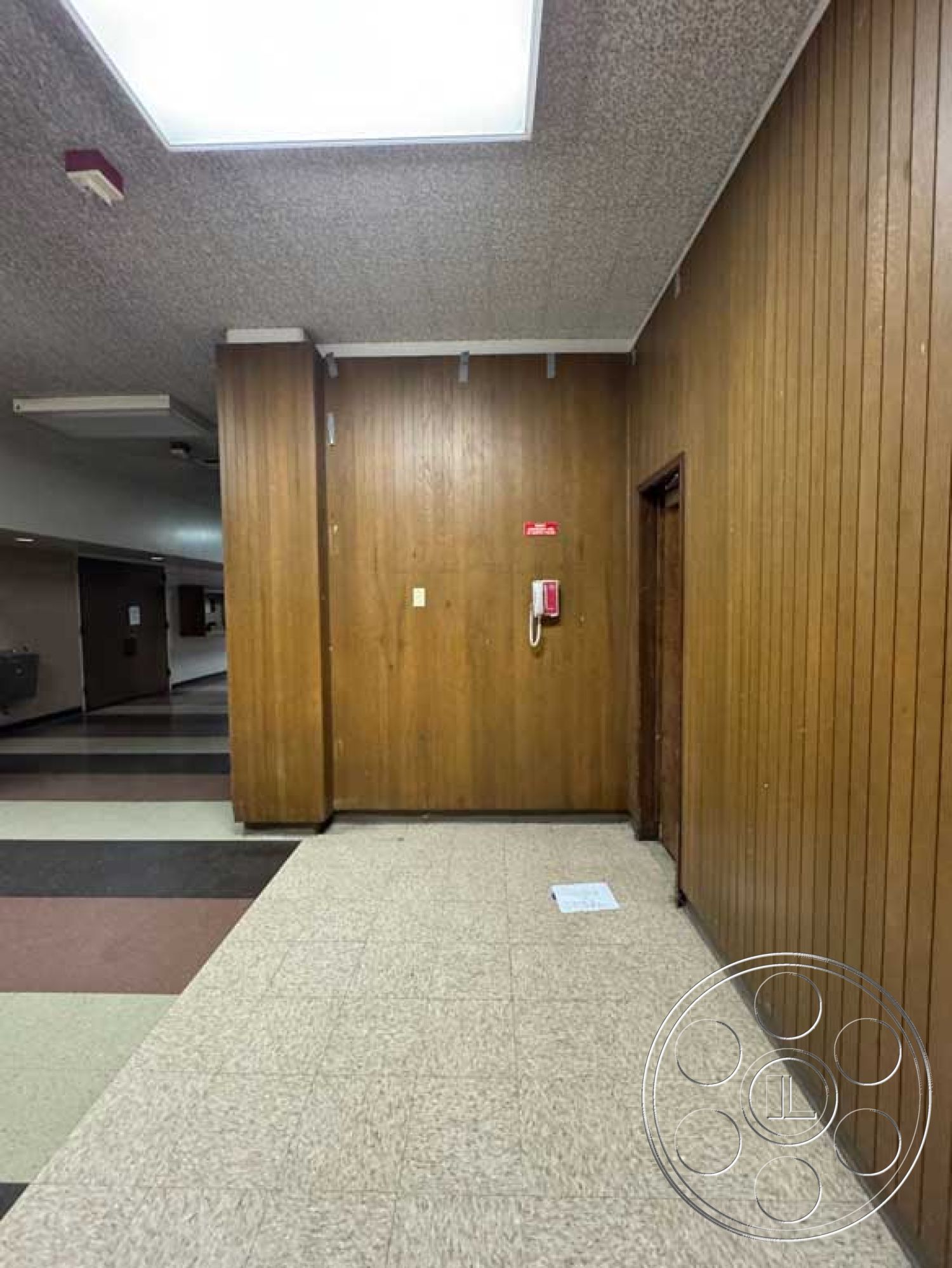 Old School - exposed light fixtures, neutral color scheme, dark wood door frame, interior wooden paneling wall, textured plaster wall details, interior acoustic ceiling tiles, tile flooring hallway, single-pane glass door, fire extinguisher wall mount, office space phone mount, rectangular wall shape, light-colored linoleum floor, vintage interior style