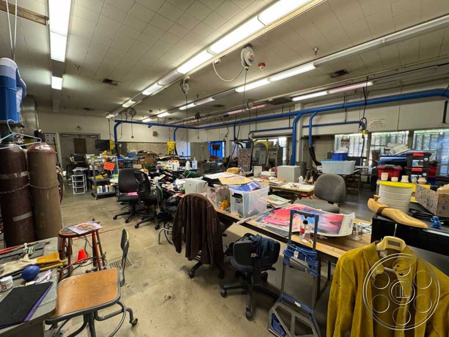 Old School - workshop environment, fluorescent lighting fixtures, interior workspace, suspended ceiling grid, rolling office chairs, plastic storage bins, commercial ceiling panels, concrete floor with stain marks, industrial metal storage cabinets, wooden tables with scattered materials, various craft supplies on tables, industrial-style workbenches, exposed ductwork and pipes, workspace clutter, paint-splattered surfaces, multiple power outlets, vertical shelving units, fabric samples and tools, creative studio layout, mixed-use design space